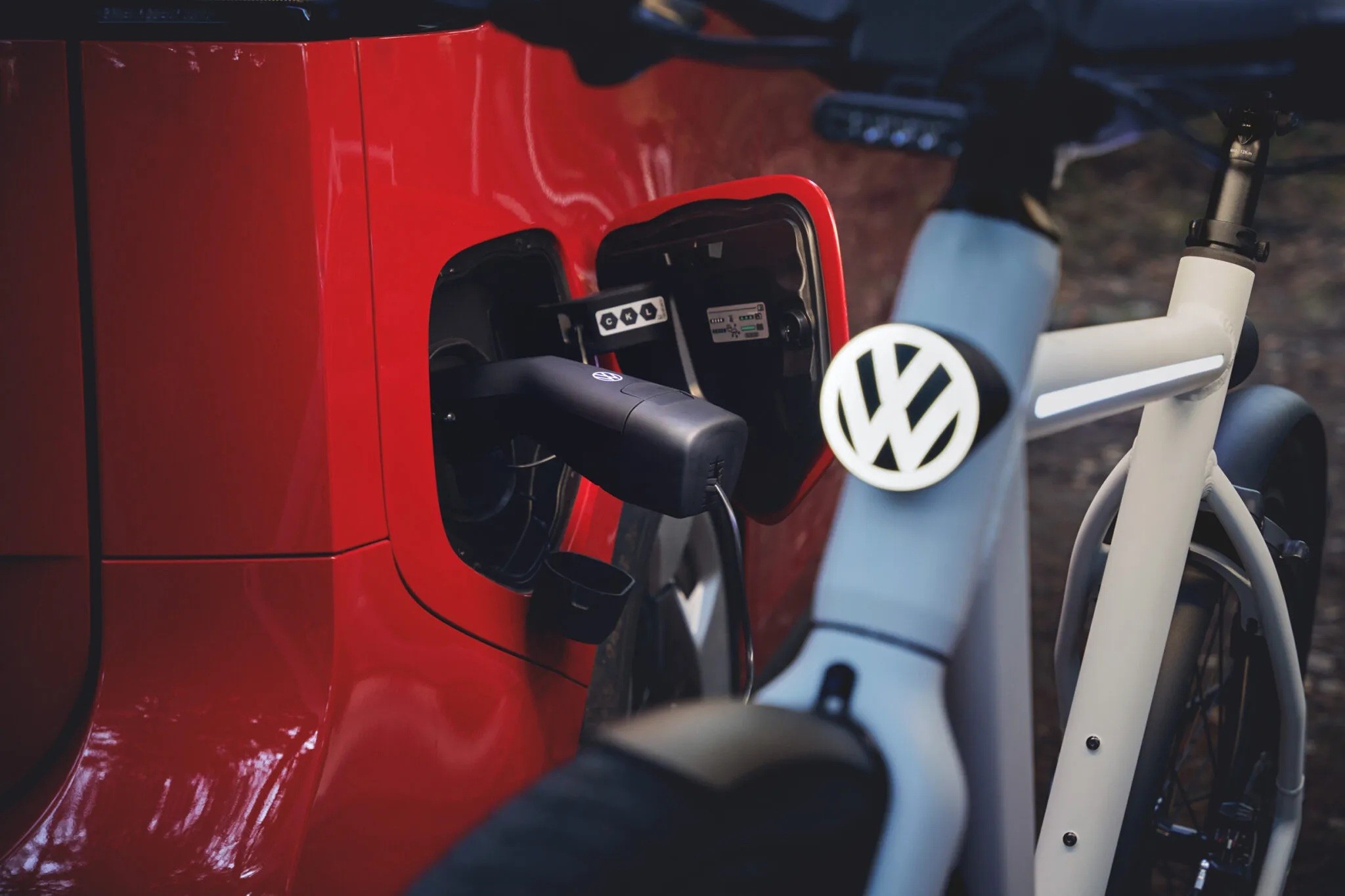 Carro vermelho sendo carregado por um cabo preto conectado à porta de carregamento, com uma bicicleta branca com o logo da Volkswagen em primeiro plano