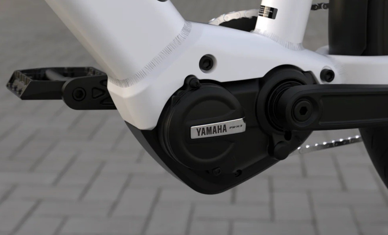Motor elétrico Yamaha preto com logo prateado, integrado ao quadro branco de uma bicicleta, com pedal preto à esquerda, sobre piso de blocos cinzas