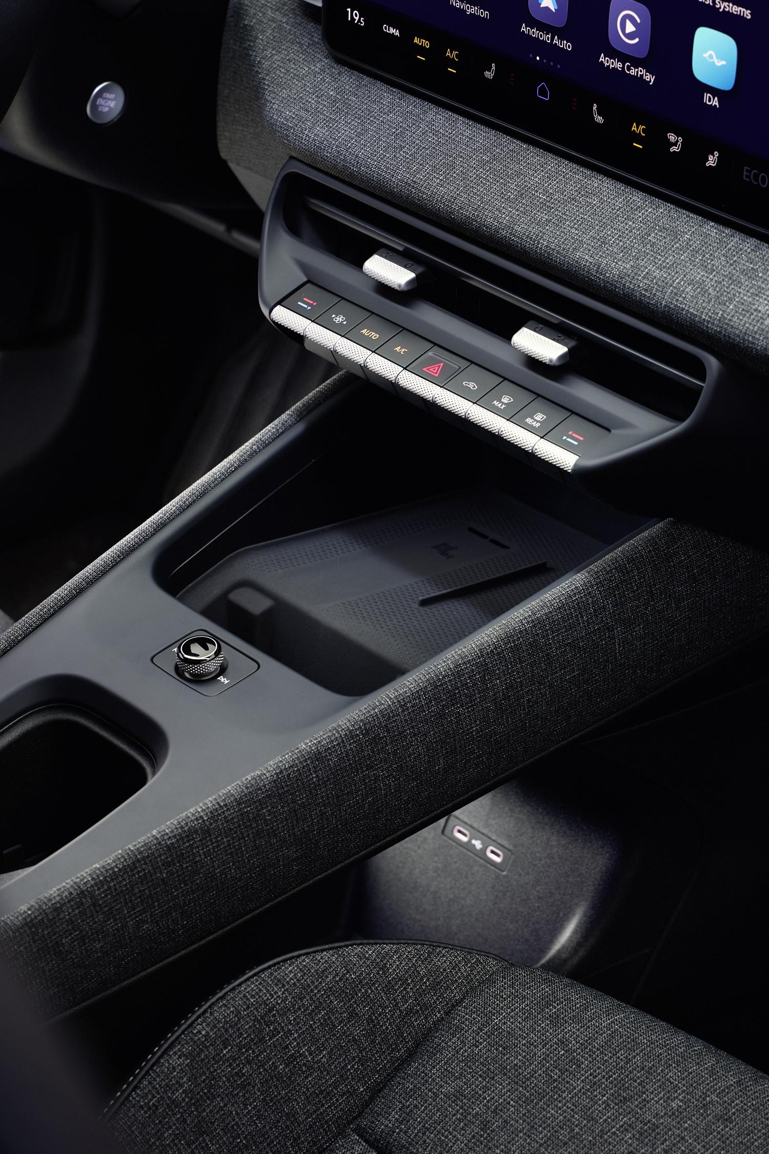 Interior de um carro moderno com painel cinza-escuro texturizado, tela multimídia exibindo Android Auto e Apple CarPlay, controles de ar-condicionado e um carregador de celular por indução no console central