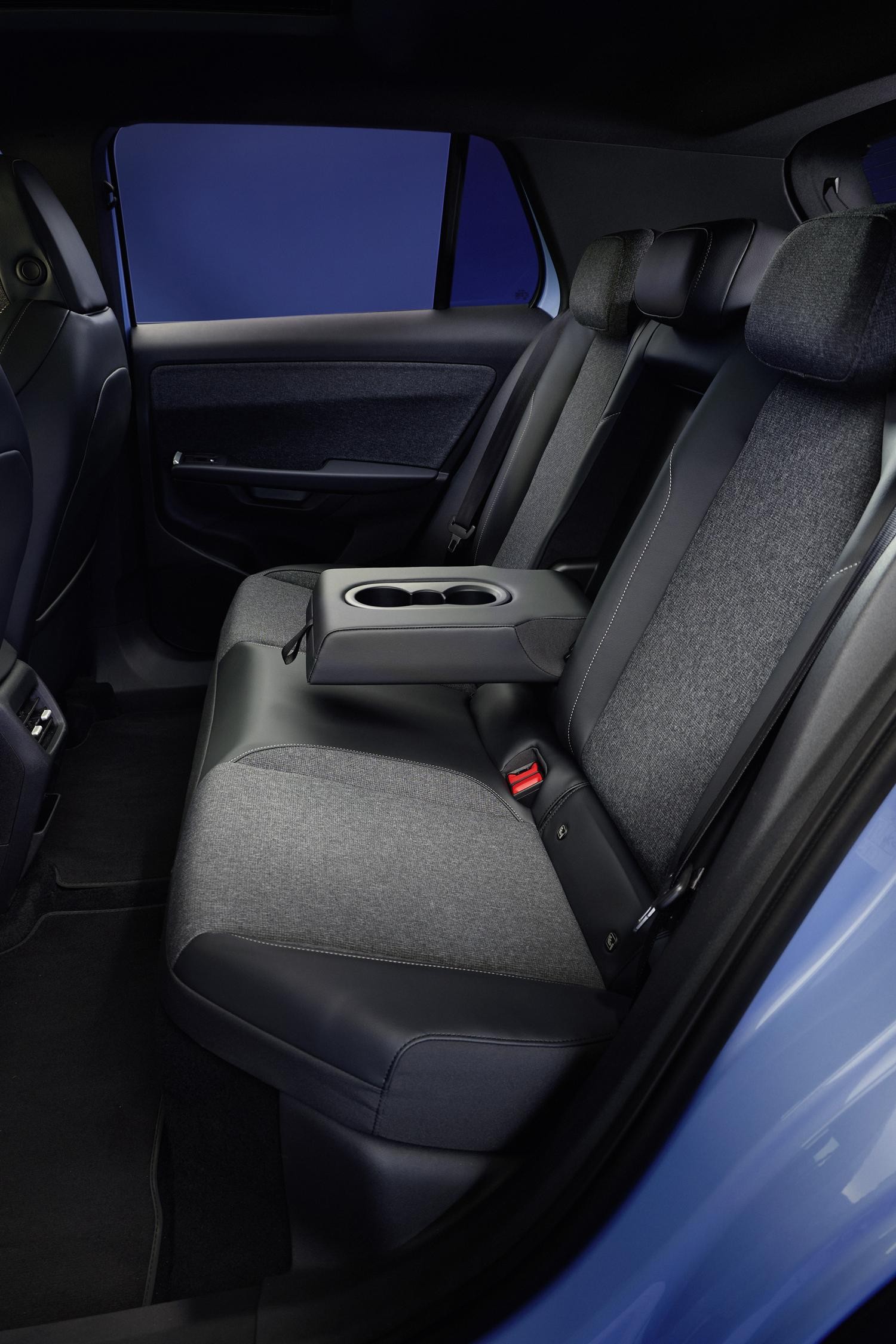 Interior de um carro com bancos traseiros em tecido cinza e detalhes em couro preto, com um apoio de braço central rebatível com porta-copos. O assoalho é preto e a janela exibe um tom azul escuro