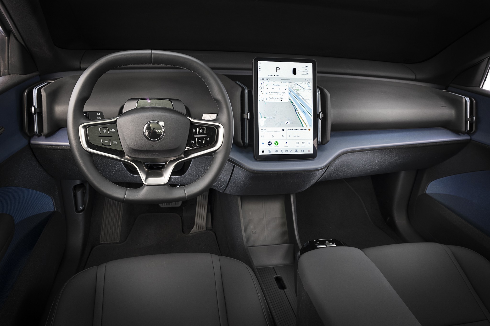 Interior de um carro moderno com volante preto e cinza, painel azul-acinzentado e tela vertical exibindo um mapa de navegação. Os bancos dianteiros são pretos, e o console central é discreto, com um apoio de braço. O ambiente é minimalista e tecnológico