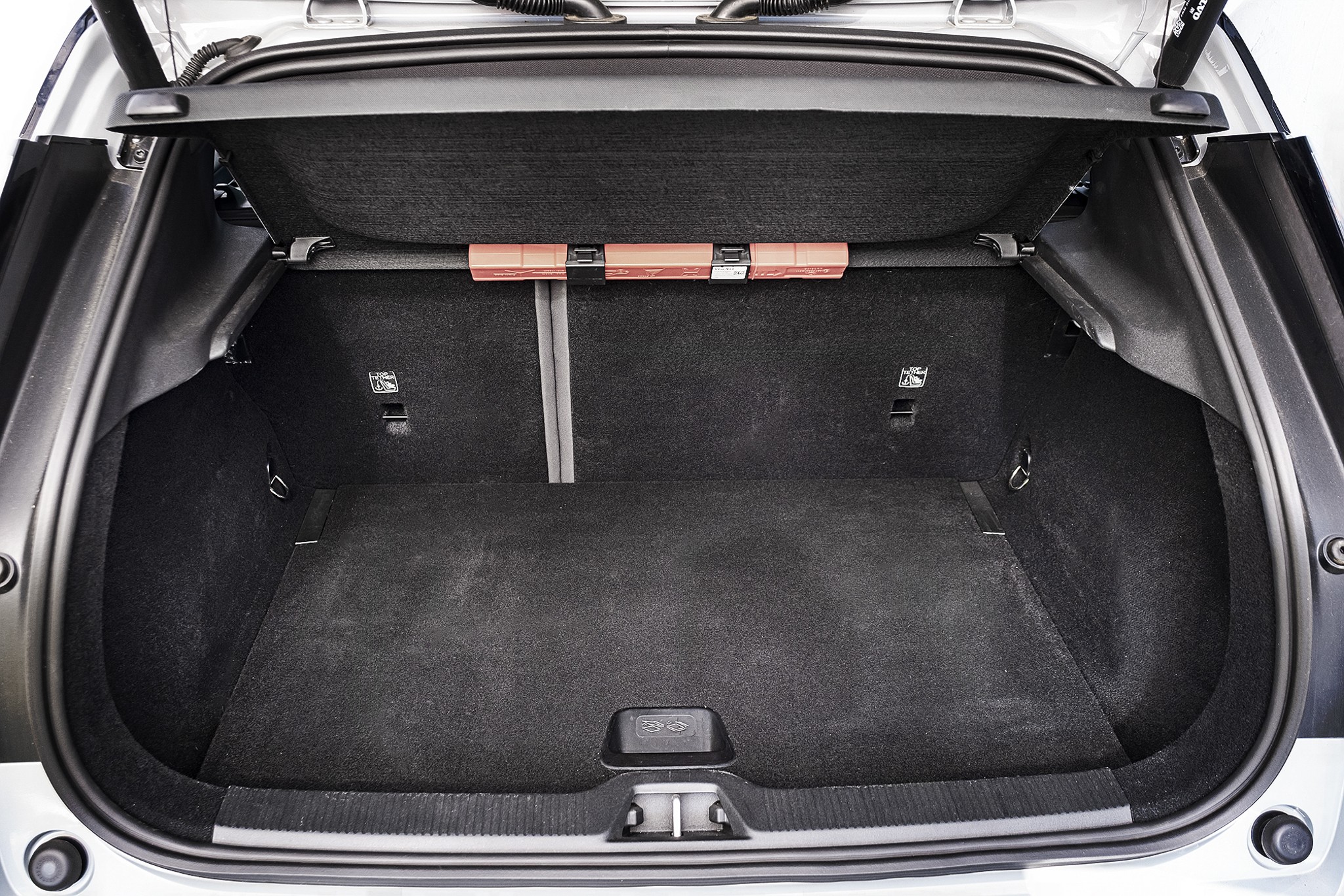 Porta-malas de carro hatch aberto, forrado com carpete cinza escuro, sem objetos. Há uma barra transversal com três refletores laranja na parte superior e ganchos laterais para fixação de carga