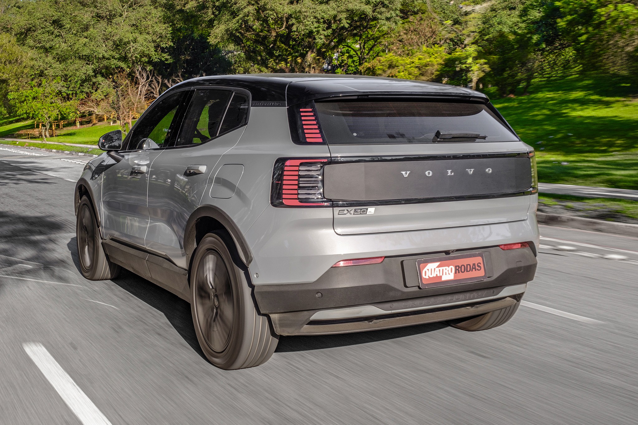 SUV Volvo EX30 cinza claro com teto preto, visto de traseira e lateral, em movimento numa estrada asfaltada com árvores verdes ao fundo
