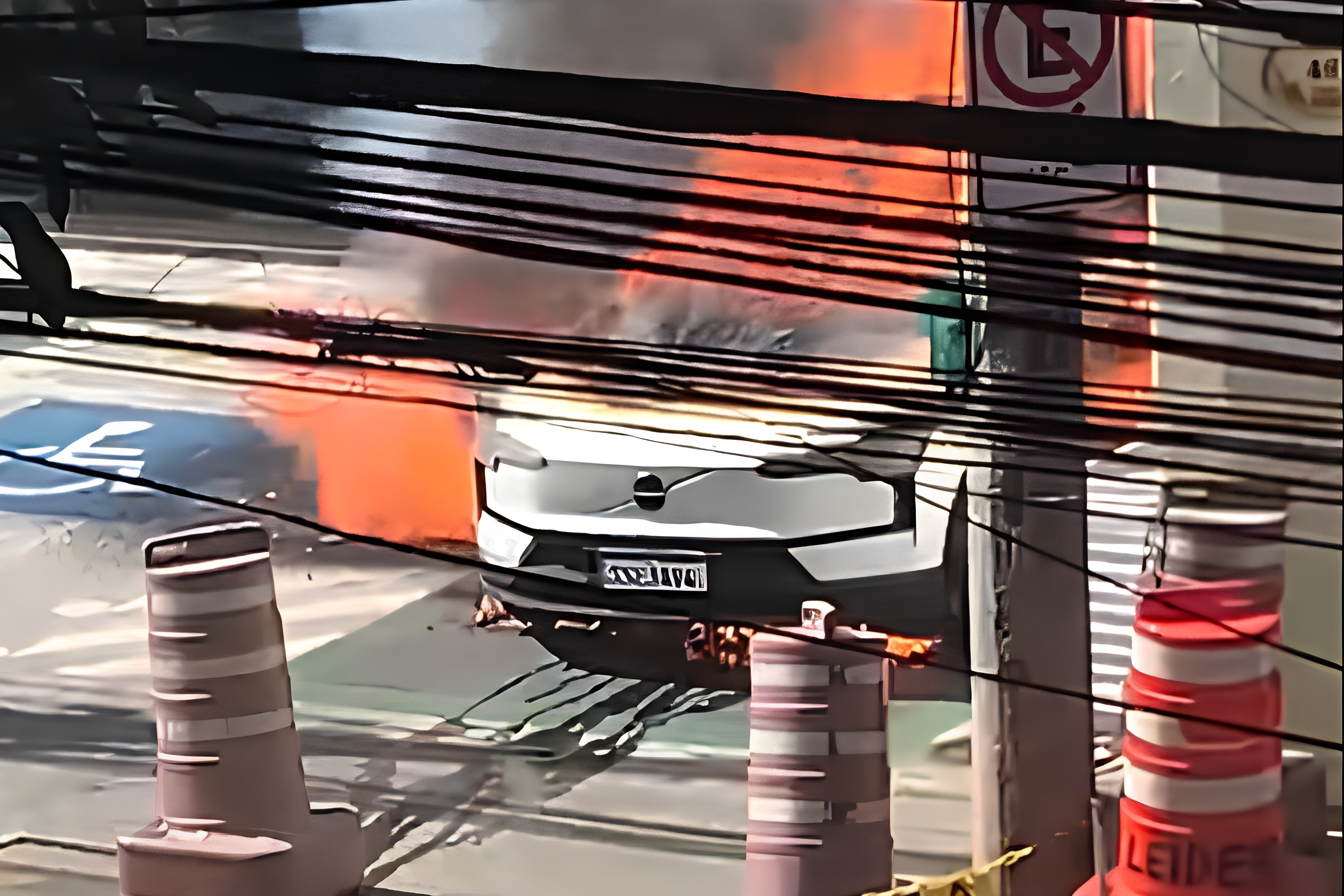 Carro branco em chamas na rua, com fumaça laranja e preta subindo. Fios elétricos cruzam a imagem em primeiro plano, e cones de trânsito estão visíveis. Um poste com placa de proibido estacionar aparece à direita