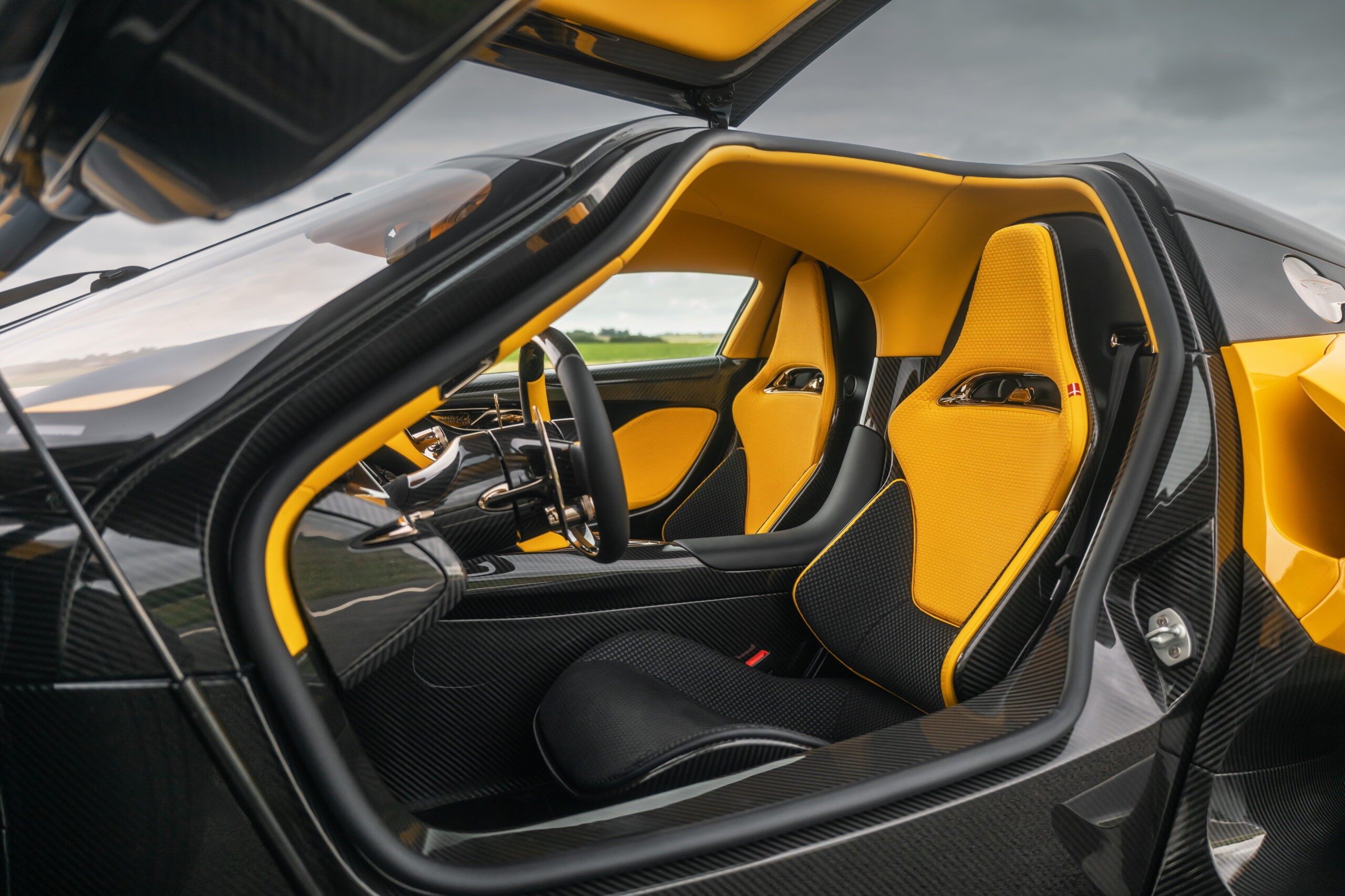 Interior de um carro esportivo de luxo, com a porta aberta revelando bancos esportivos pretos e amarelos, painel e volante pretos com detalhes amarelos, e acabamento em fibra de carbono. O teto também é amarelo.