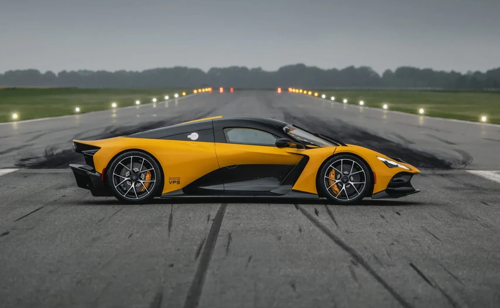 Carro esportivo amarelo e preto, perfil lateral, parado em uma pista de asfalto com luzes de balizamento ao fundo, sob céu nublado