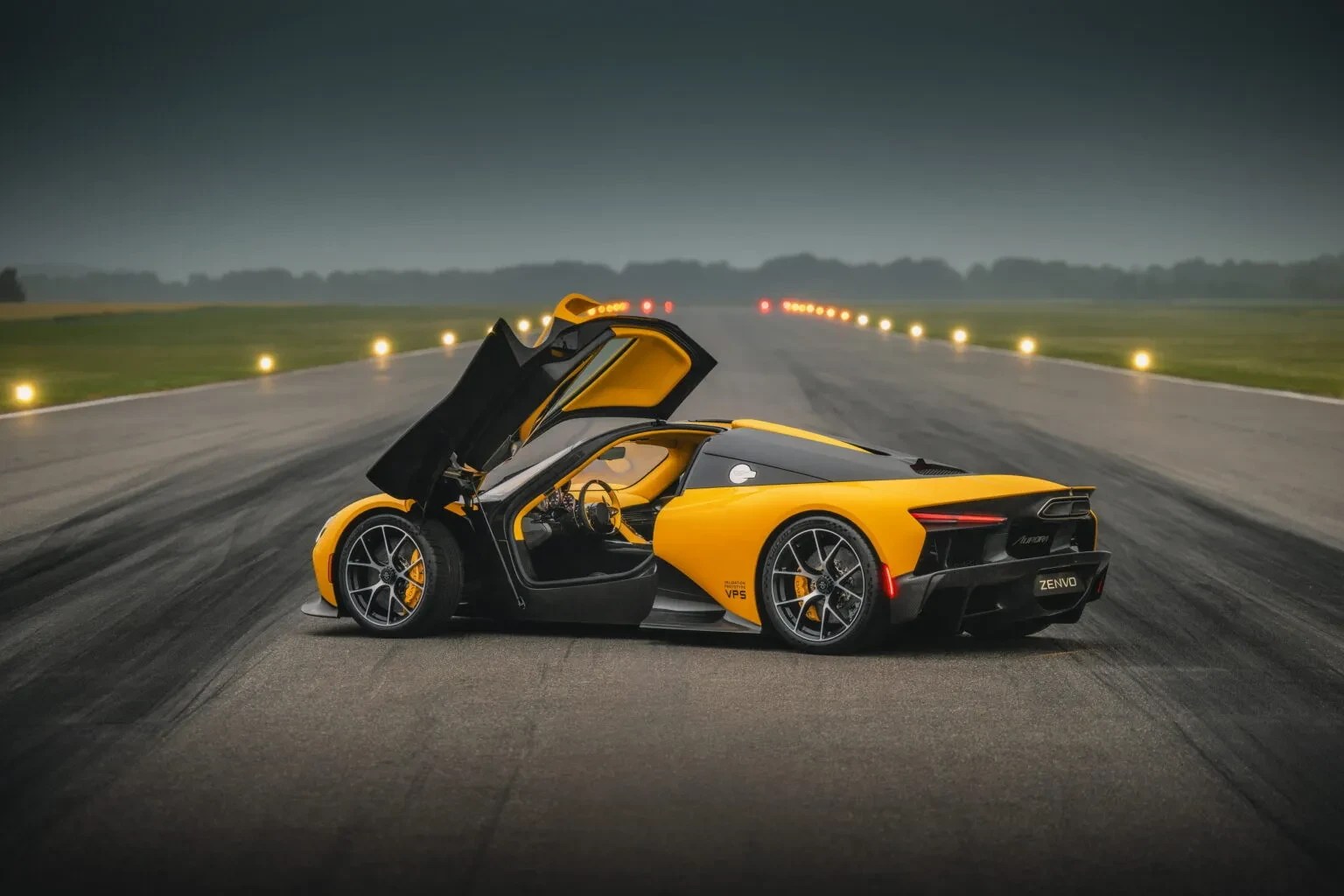 Carro esportivo Zenvo Aurora amarelo e preto com portas abertas para cima, parado em uma pista de asfalto com luzes de balizamento ao fundo sob um céu nublado