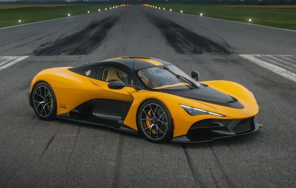 Carro esportivo amarelo e preto, com design futurista, parado em uma pista de asfalto molhada. Ao fundo, uma pista de pouso com luzes e um céu nublado e escuro.