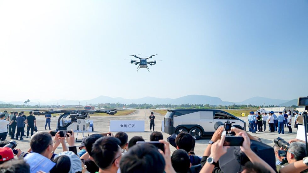 Um drone de oito hélices paira no ar, observado por uma multidão em um evento ao ar livre. Pessoas filmam com celulares, e um veículo futurista branco está à direita. Montanhas distantes e céu azul claro compõem o cenário