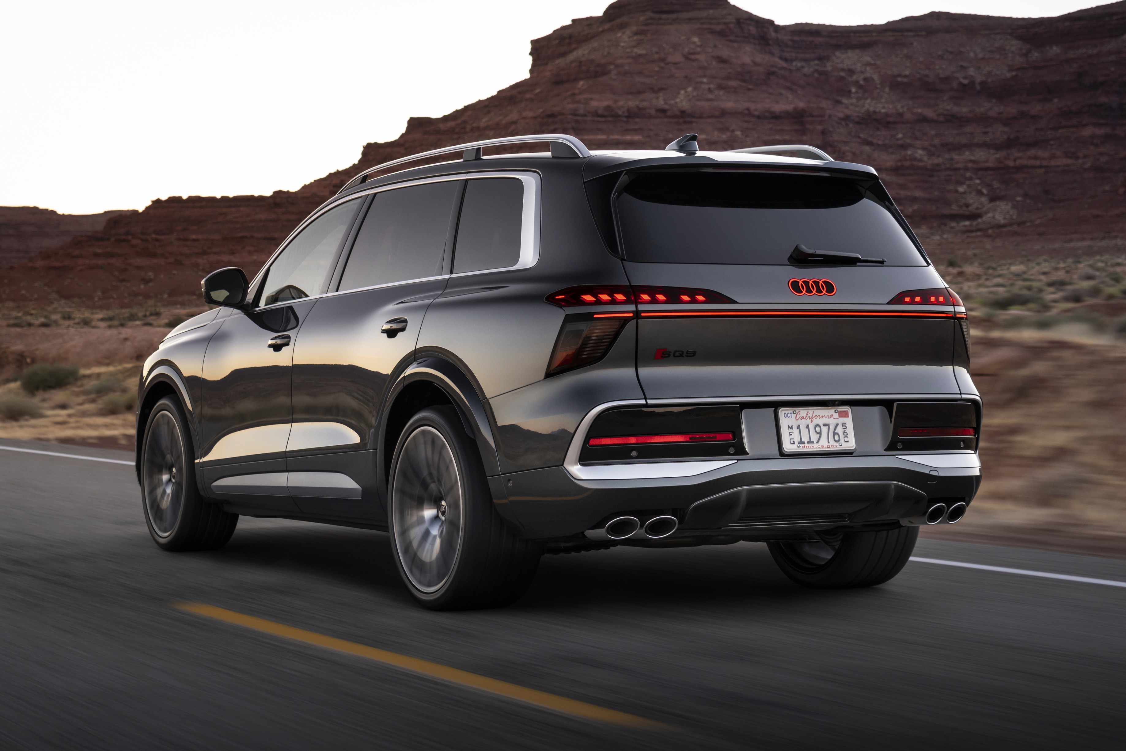 SUV Audi Q9 cinza escuro em movimento, visto por trás e lateralmente, em uma estrada com linha amarela. Ao fundo, montanhas rochosas e céu claro.