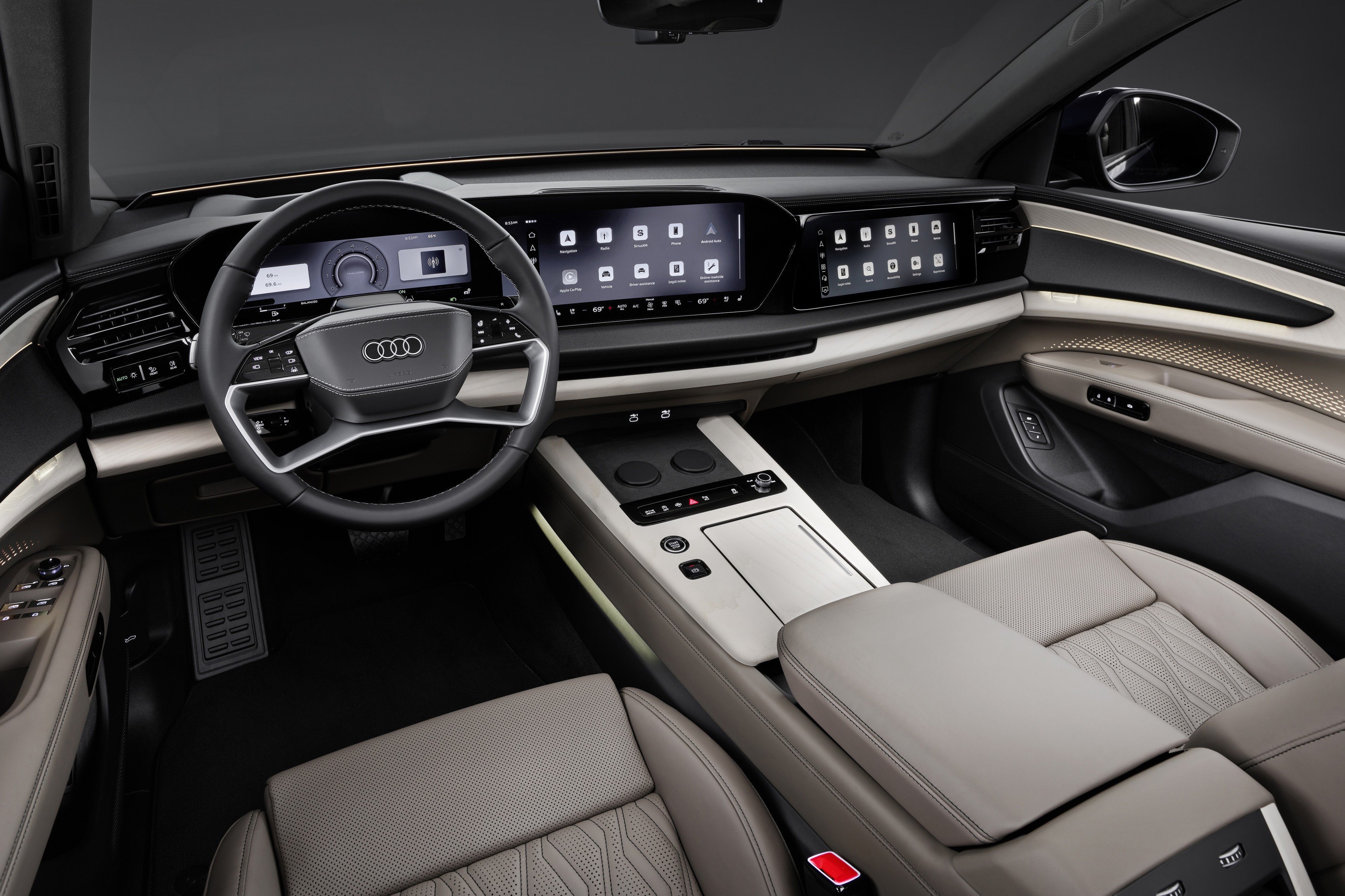 Interior de um carro Audi moderno, com volante multifuncional preto e o logotipo da marca. O painel possui três telas digitais, exibindo ícones de aplicativos. O console central é cinza claro, com botões e porta-copos. Os bancos são de couro cinza, com detalhes texturizados. A porta do passageiro tem acabamento em cinza e bege, com detalhes em malha dourada. O ambiente é sofisticado e tecnológico