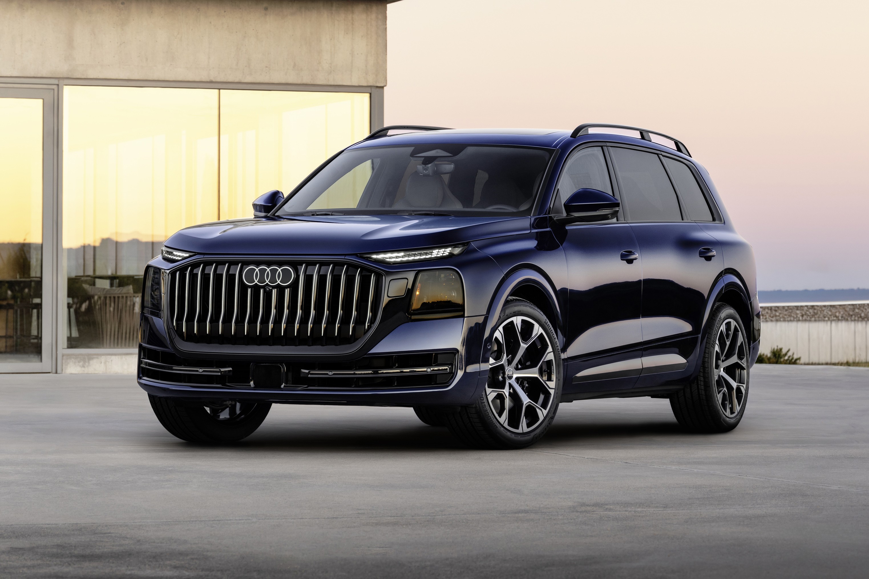 SUV Audi Q9 azul-escuro estacionado em uma área externa de concreto, com uma construção moderna de janelas grandes e claras ao fundo. O céu está em tons de rosa e azul no horizonte, e há uma faixa de água visível à direita