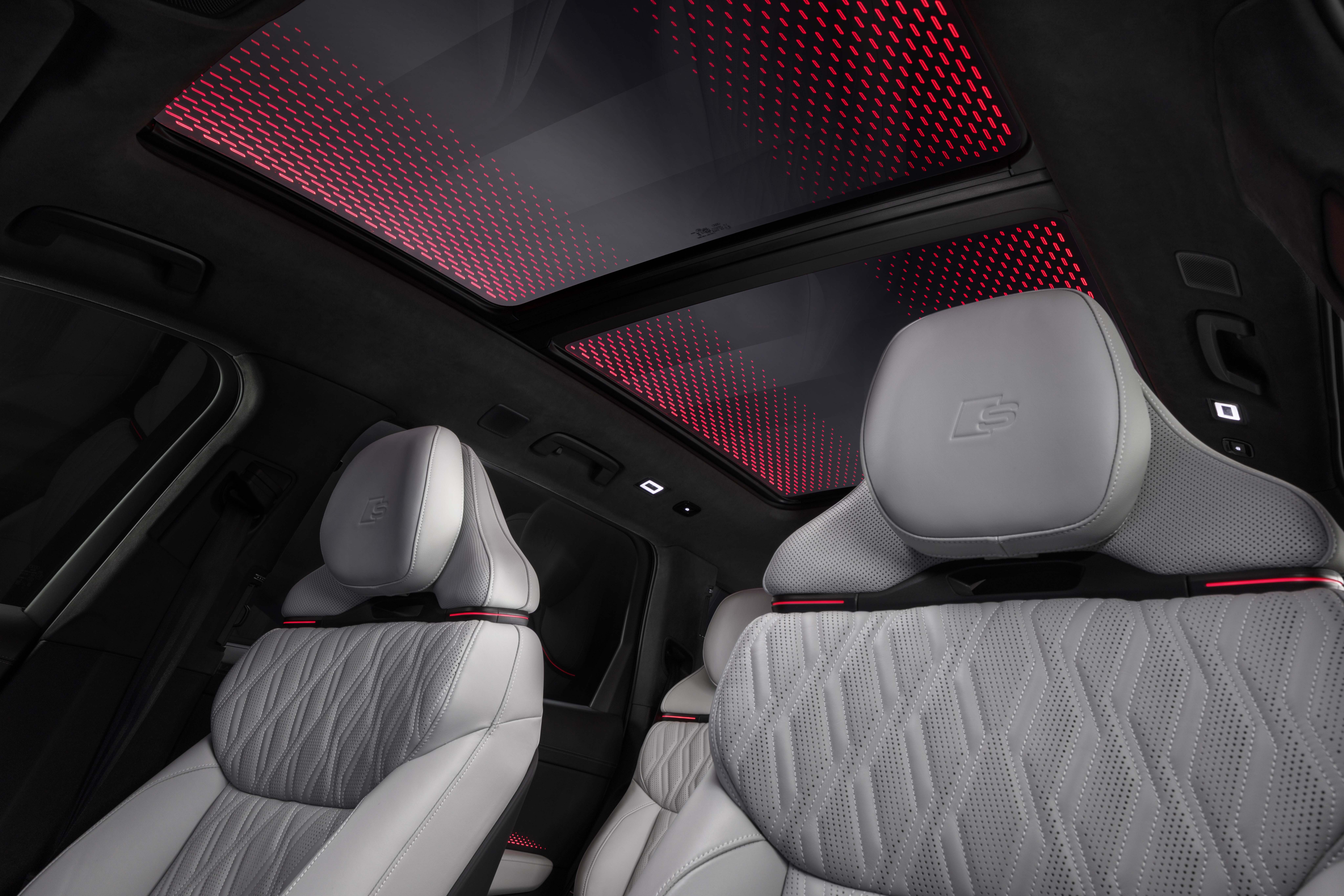Interior de carro luxuoso com bancos de couro cinza claro e costura em losango, encostos de cabeça com logo. Teto solar panorâmico exibe iluminação ambiente vermelha com padrão geométrico