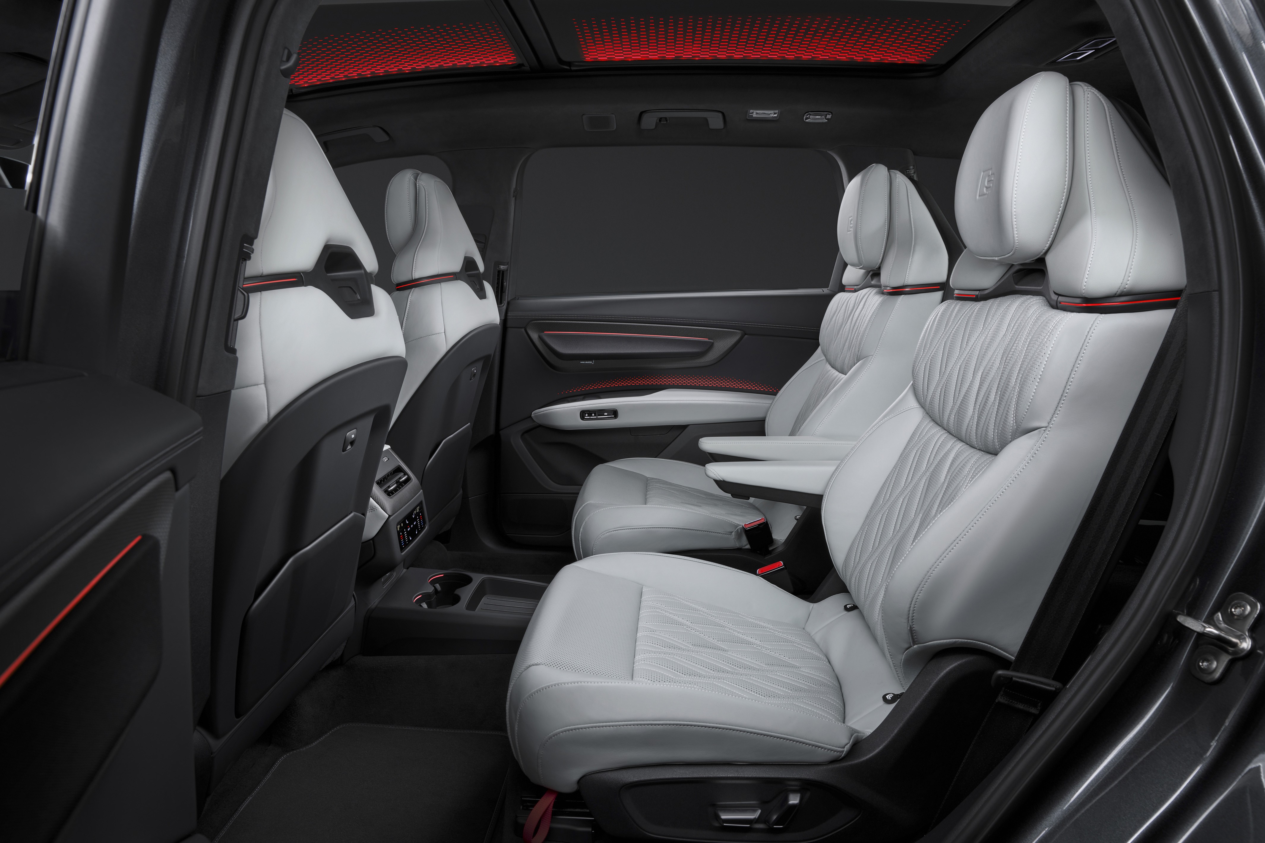 Interior de um carro de luxo com bancos de couro branco e detalhes em vermelho, incluindo o teto solar iluminado em vermelho. Os bancos traseiros são individuais, com apoios de braço e encostos de cabeça ajustáveis, e há um console central com porta-copos. As portas têm acabamento escuro com frisos vermelhos, e o ambiente é sofisticado