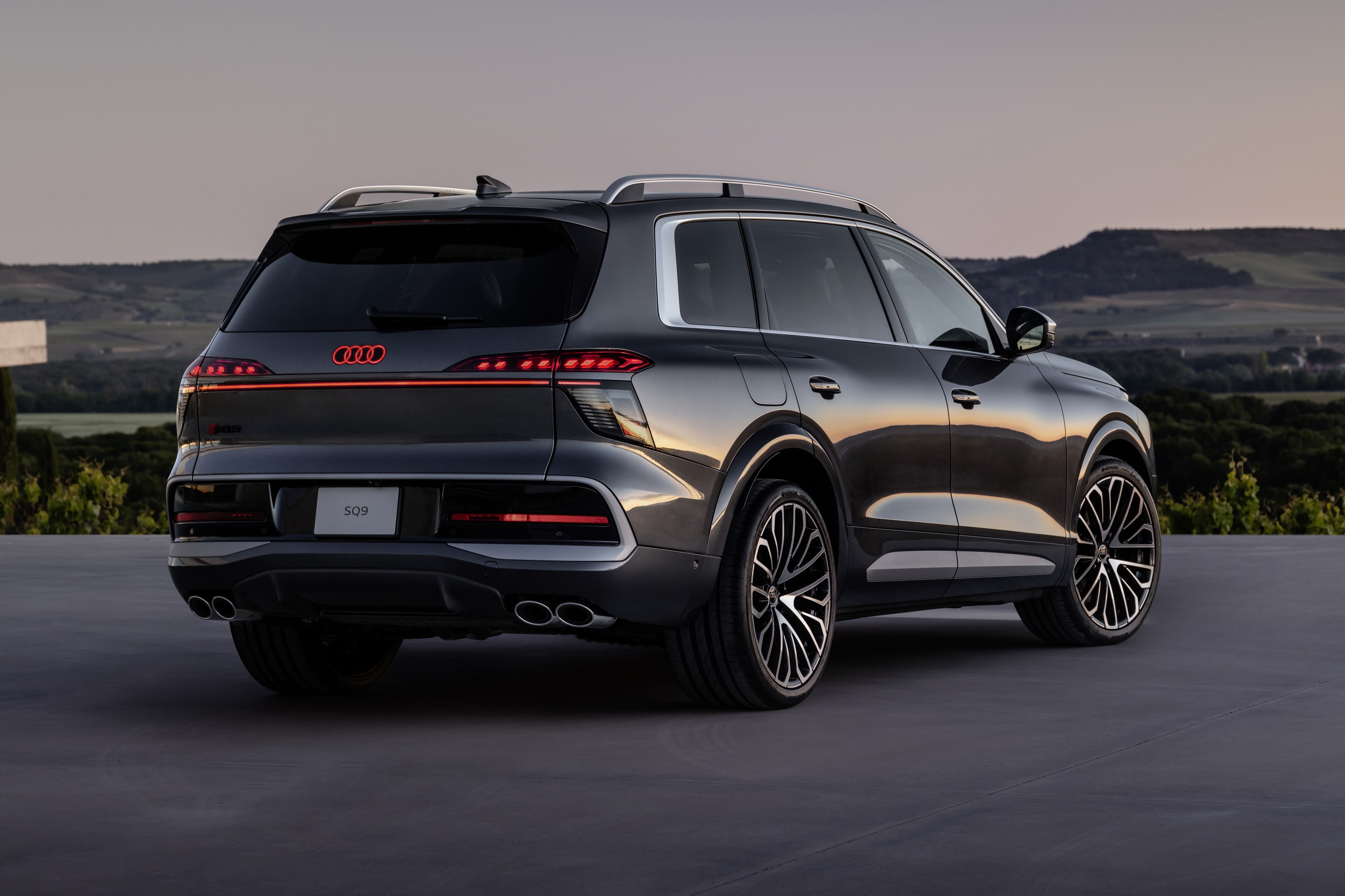 SUV Audi Q9 cinza escuro, visto por trás e levemente de lado, com lanternas traseiras vermelhas acesas e o logotipo Audi em vermelho. O carro está estacionado em uma superfície escura, com um cenário de montanhas e vegetação ao fundo sob um céu claro