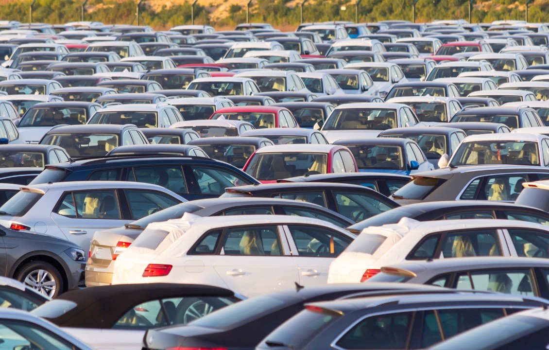 Estacionamento lotado com centenas de carros novos e usados, de diversas cores como branco, cinza, azul e vermelho, sob a luz do sol
