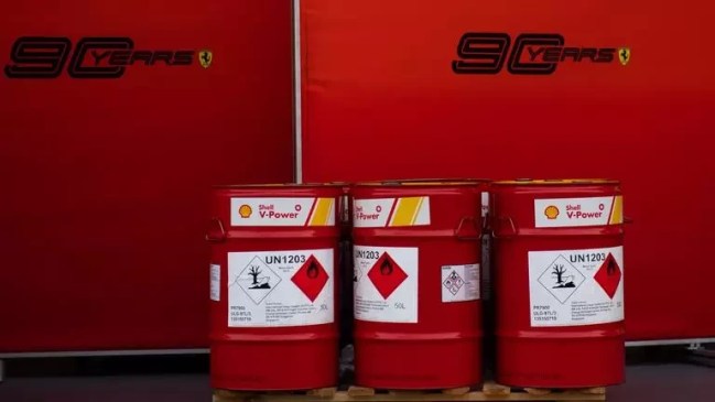 Três tambores vermelhos de 50 litros de combustível Shell V-Power, com rótulos de perigo de inflamabilidade e risco ambiental, sobre um palete de madeira. Ao fundo, um painel vermelho com o logo 90 Years e o emblema da Ferrari