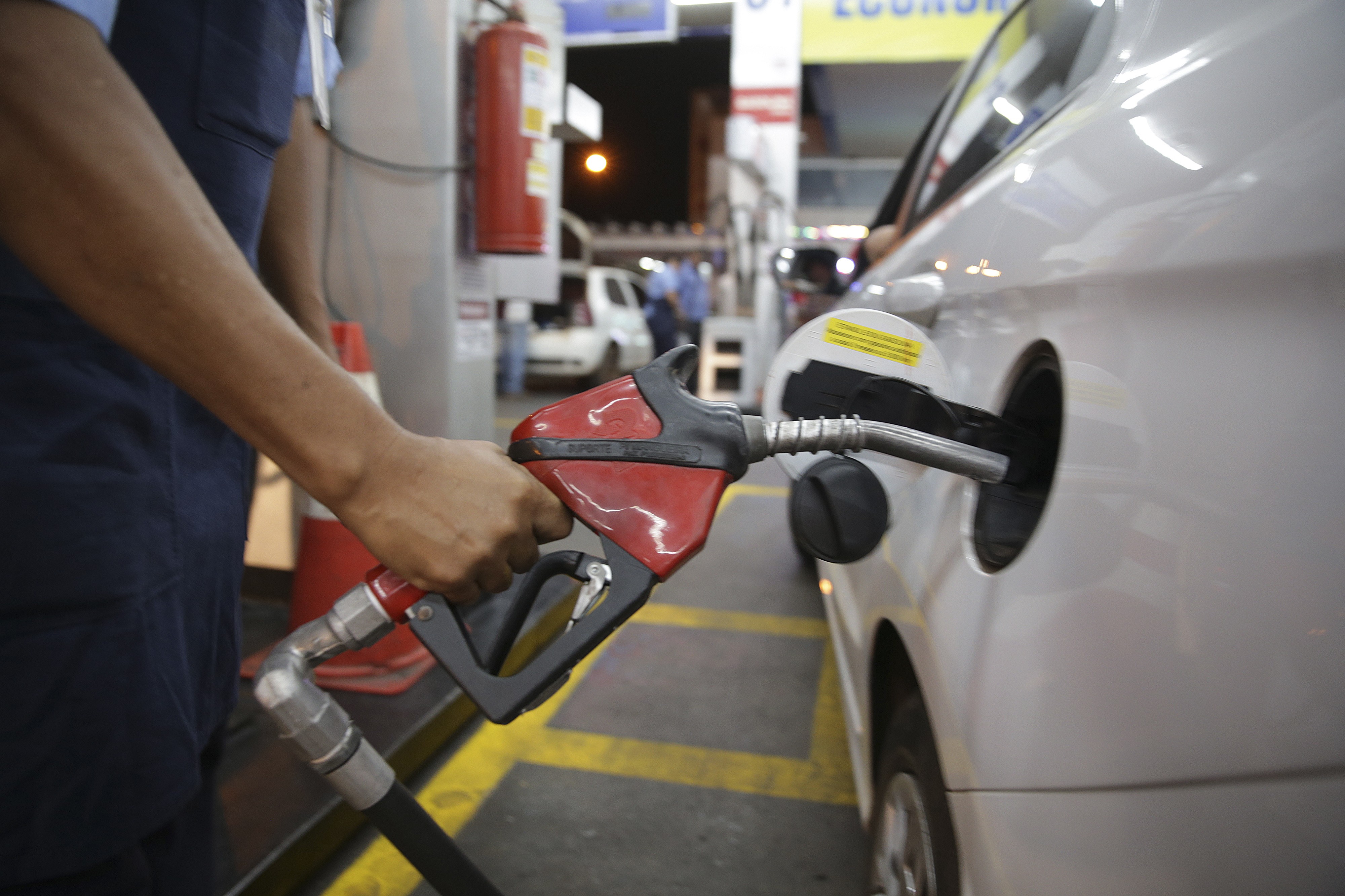 Mão de um frentista segurando um bico de bomba de combustível vermelho e preto, inserido no tanque de um carro branco em um posto de gasolina à noite. Ao fundo, um extintor de incêndio vermelho e outras bombas de combustível