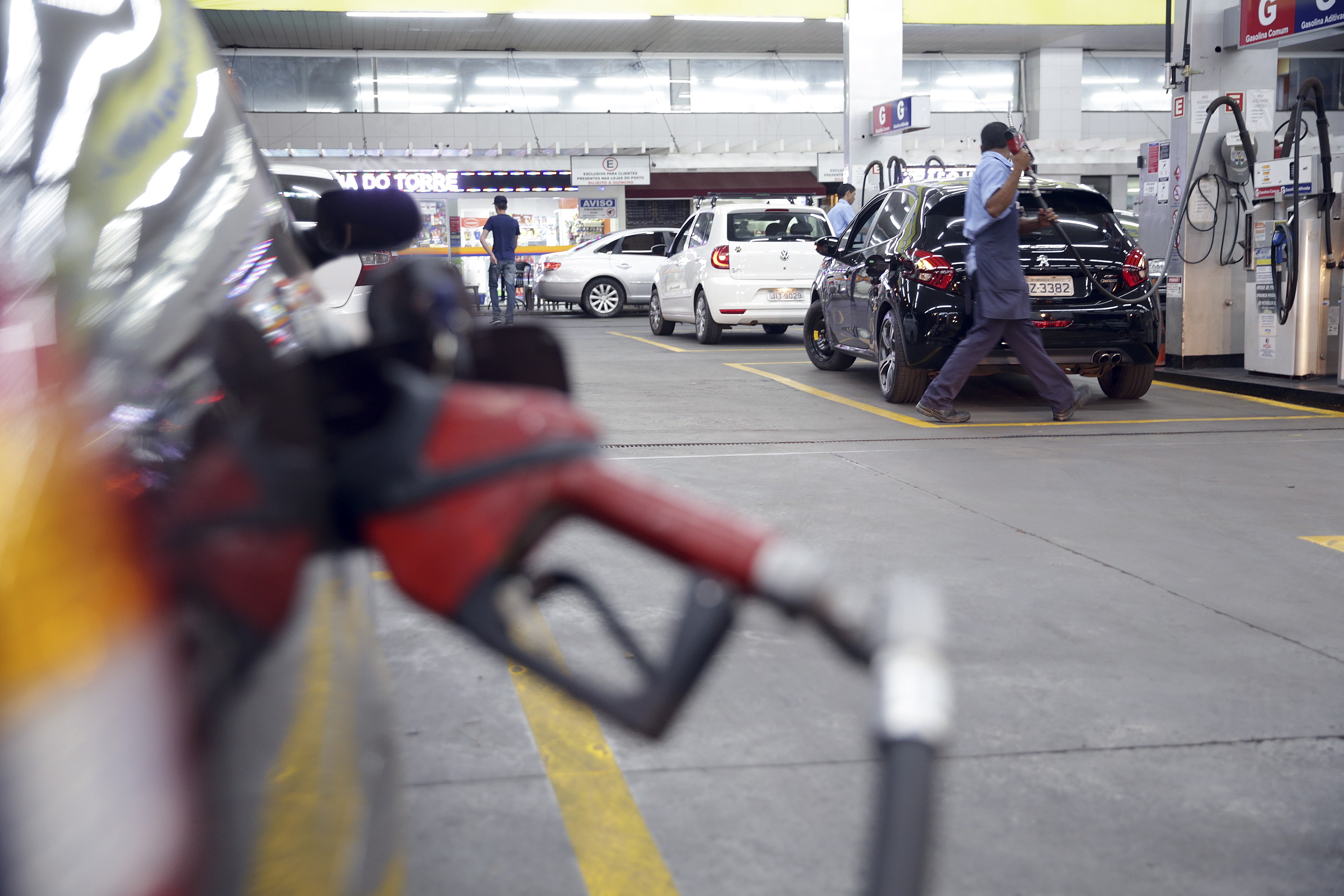 Bico de bomba de combustível vermelho em primeiro plano, desfocado. Ao fundo, um frentista de camisa azul e avental escuro abastece um carro preto em um posto de gasolina movimentado, com outros veículos e pessoas aguardando