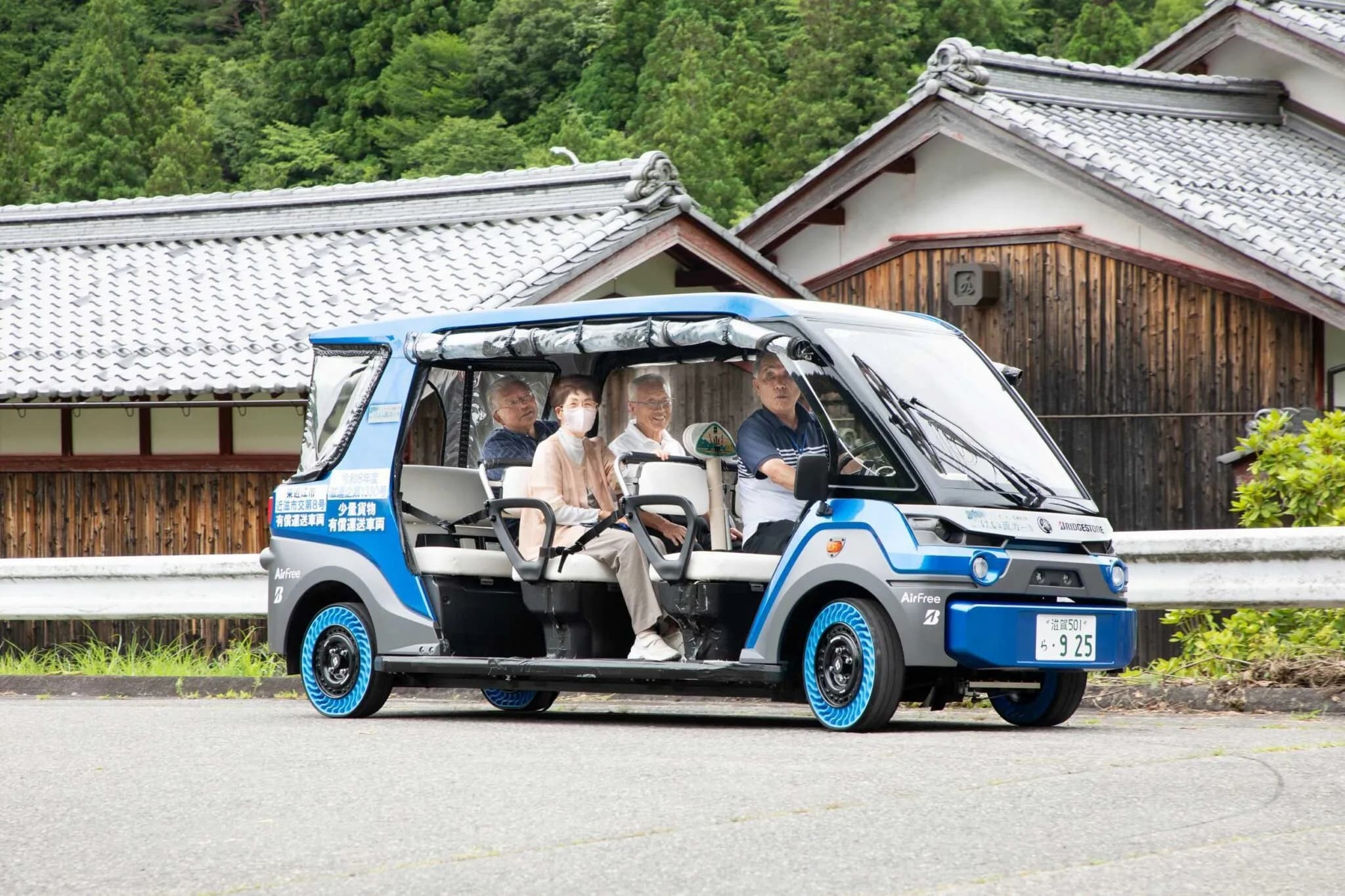 Quatro pessoas idosas, tr&ecirc;s homens e uma mulher, passeiam em um ve&iacute;culo el&eacute;trico azul e cinza com pneus azuis, em uma rua asfaltada. Ao fundo, casas tradicionais japonesas com telhados de telha e &aacute;rvores verdes. A mulher usa m&aacute;scara facial