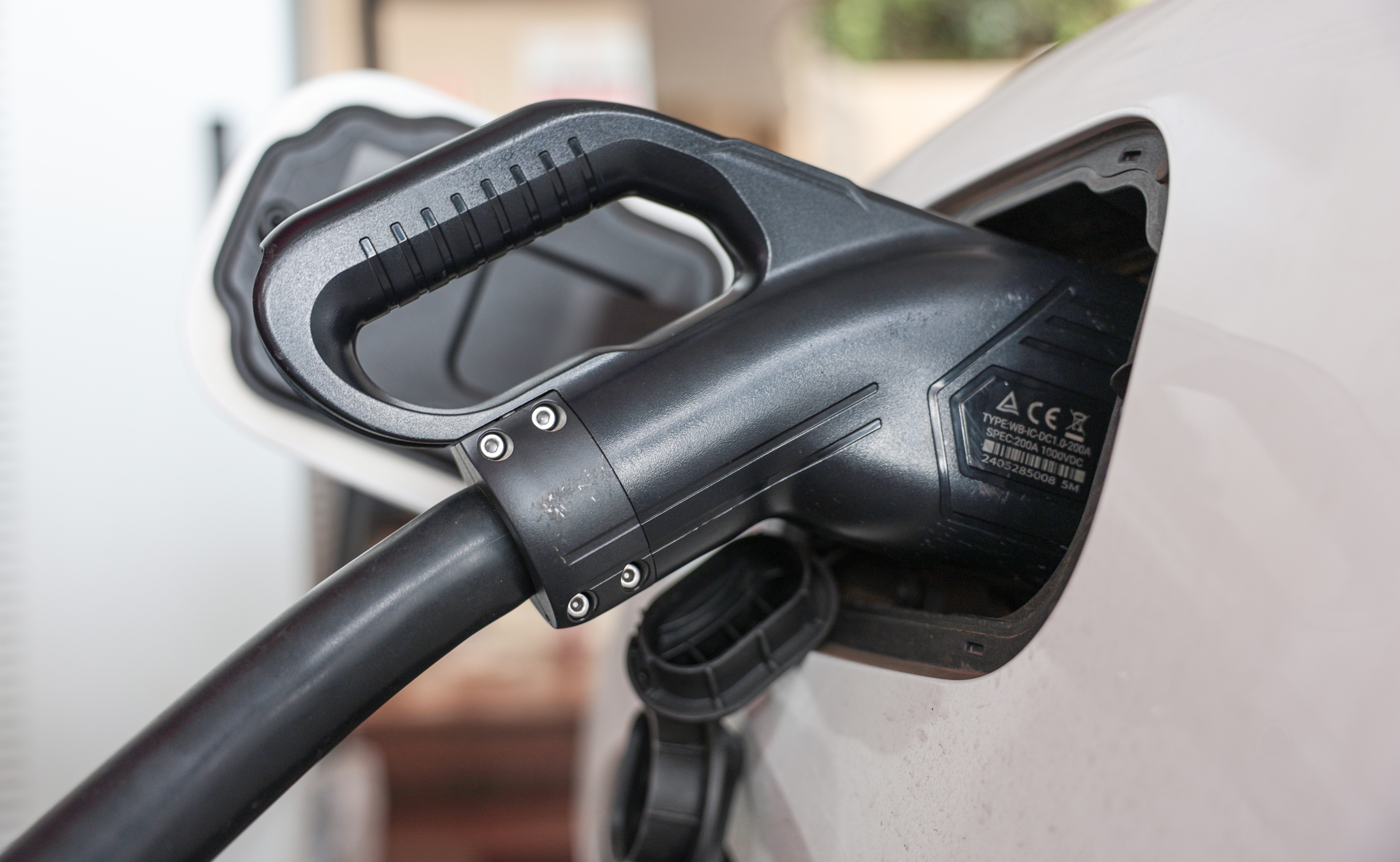 Um carregador preto de veículos elétricos conectado à porta de carregamento de um carro branco, com detalhes técnicos visíveis no carregador