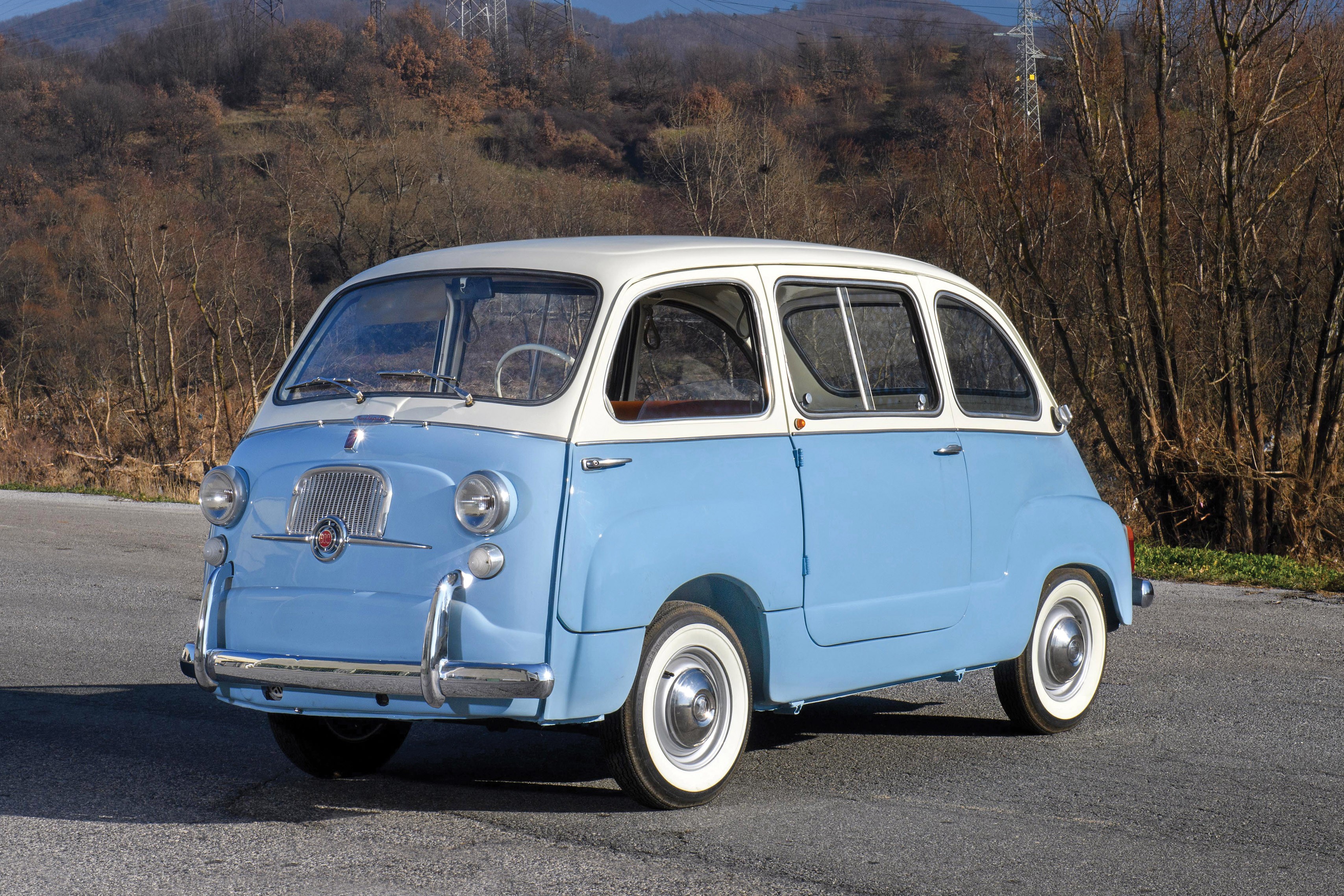 Uma van Fiat 600 Multipla azul claro e branca, estilo retrô, estacionada em uma estrada asfaltada com árvores secas e montanhas ao fundo