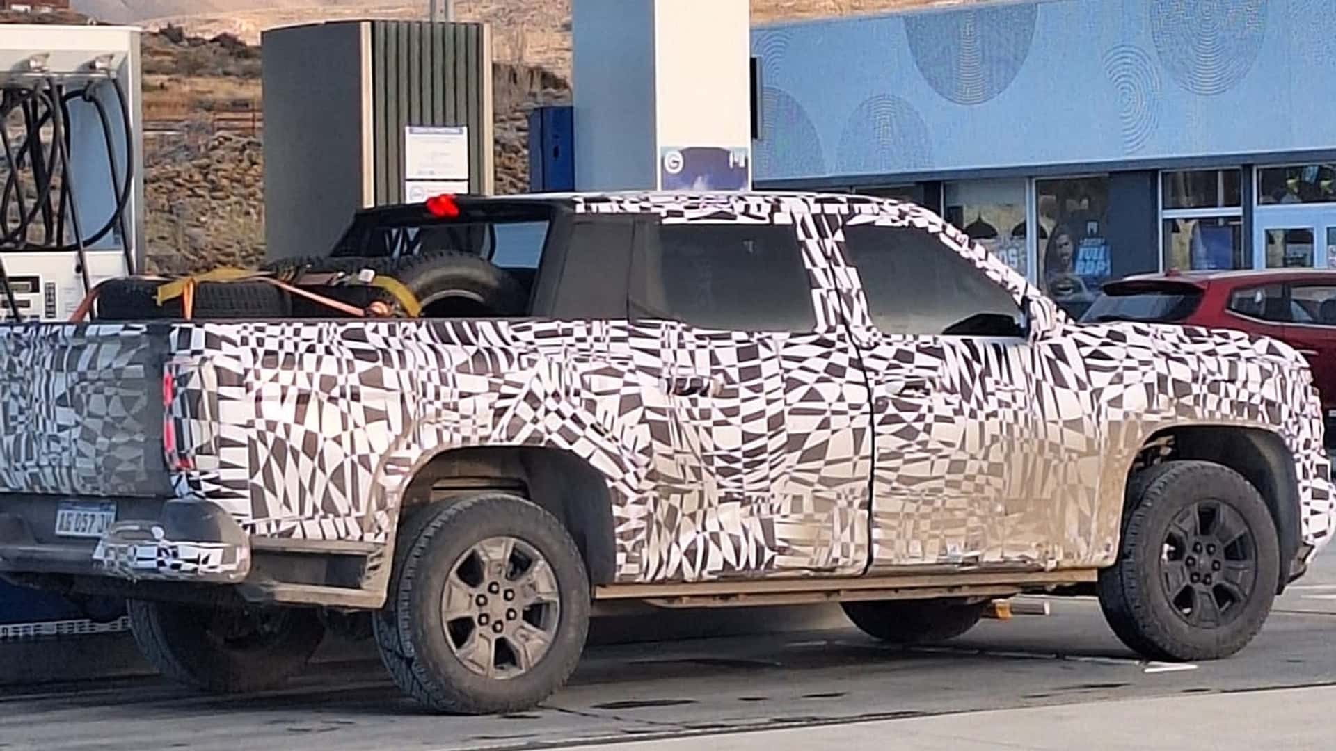 Picape camuflada com padr&atilde;o geom&eacute;trico preto e branco, pneus sobressalentes na ca&ccedil;amba, estacionada em posto de gasolina com outra picape vermelha ao fundo