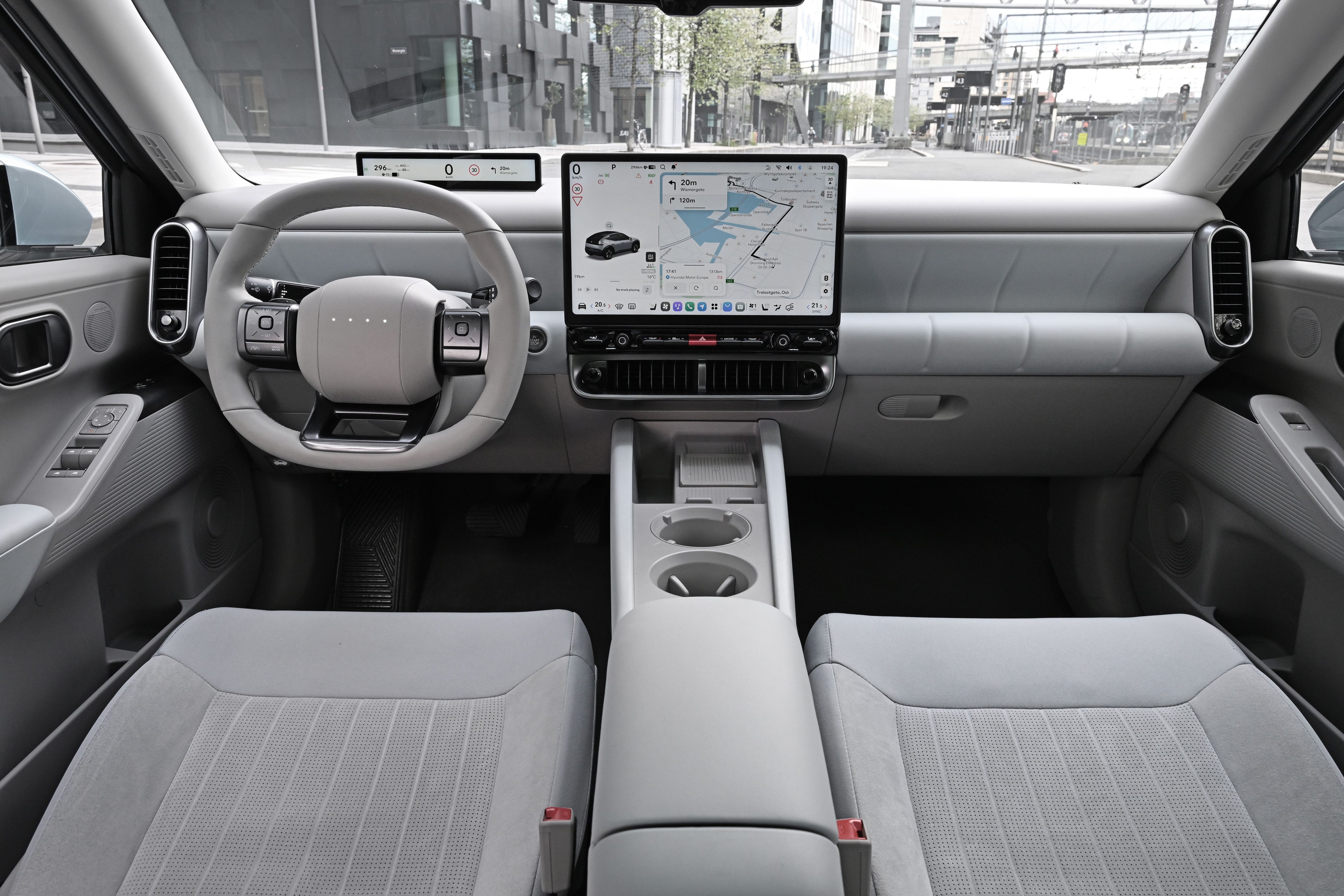 Interior de um carro moderno com acabamento cinza claro. Volante minimalista, painel digital e tela central exibindo um mapa de navegação. Bancos dianteiros estofados em tecido cinza com padrão quadriculado. Console central com porta-copos.