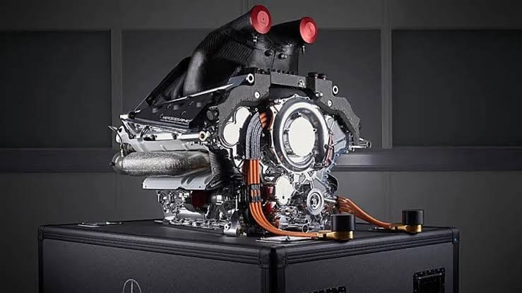 Motor de carro de corrida prateado e preto, com detalhes em vermelho e fios laranja, exibido sobre uma caixa preta em um ambiente escuro