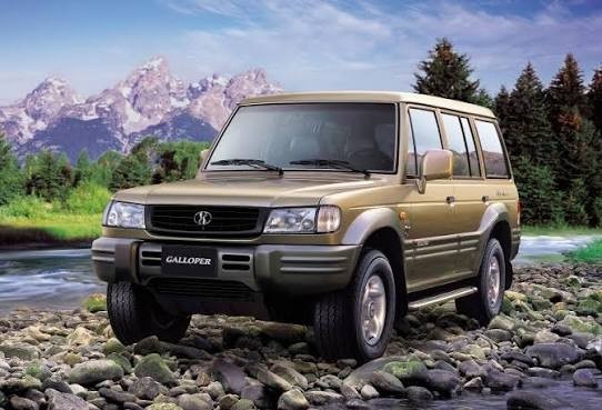 SUV Galloper dourado, com para-choques e molduras das rodas cinza-escuro, parado sobre pedras de rio. Ao fundo, montanhas nevadas, árvores e céu azul