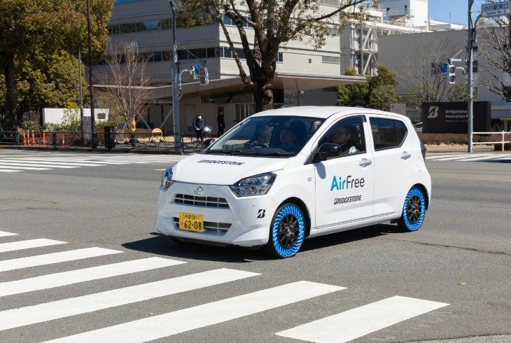 Dois carros brancos idênticos, com pneus azuis sem ar, cruzam uma faixa de pedestres em uma rua urbana ensolarada