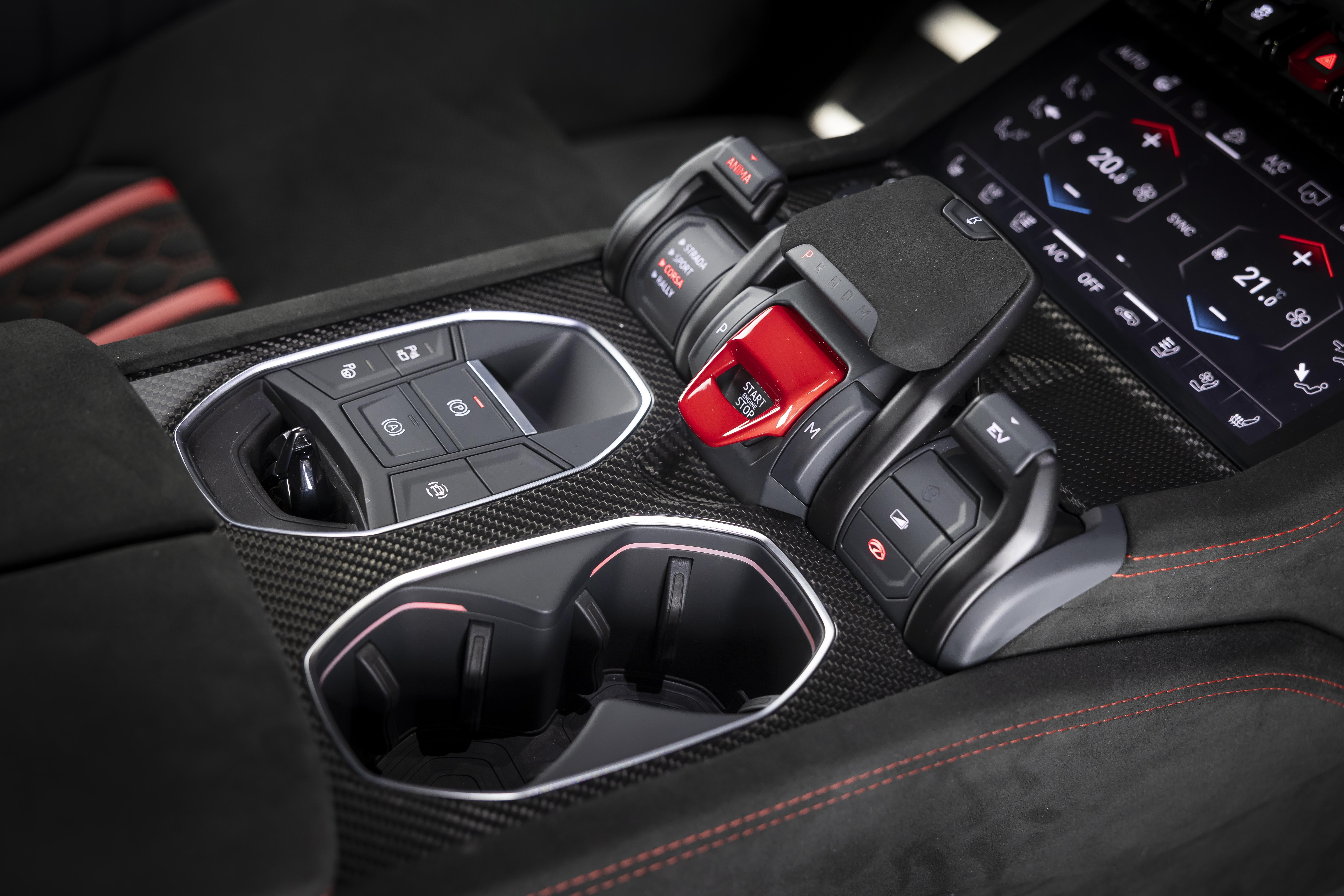 Console central de um carro esportivo, com acabamento em fibra de carbono. À esquerda, um painel de botões de controle. Ao centro, o botão vermelho de partida do motor, protegido por uma tampa. À direita, uma tela digital exibe controles de temperatura. Abaixo, dois porta-copos. O estofamento preto com costura vermelha é visível nas laterais