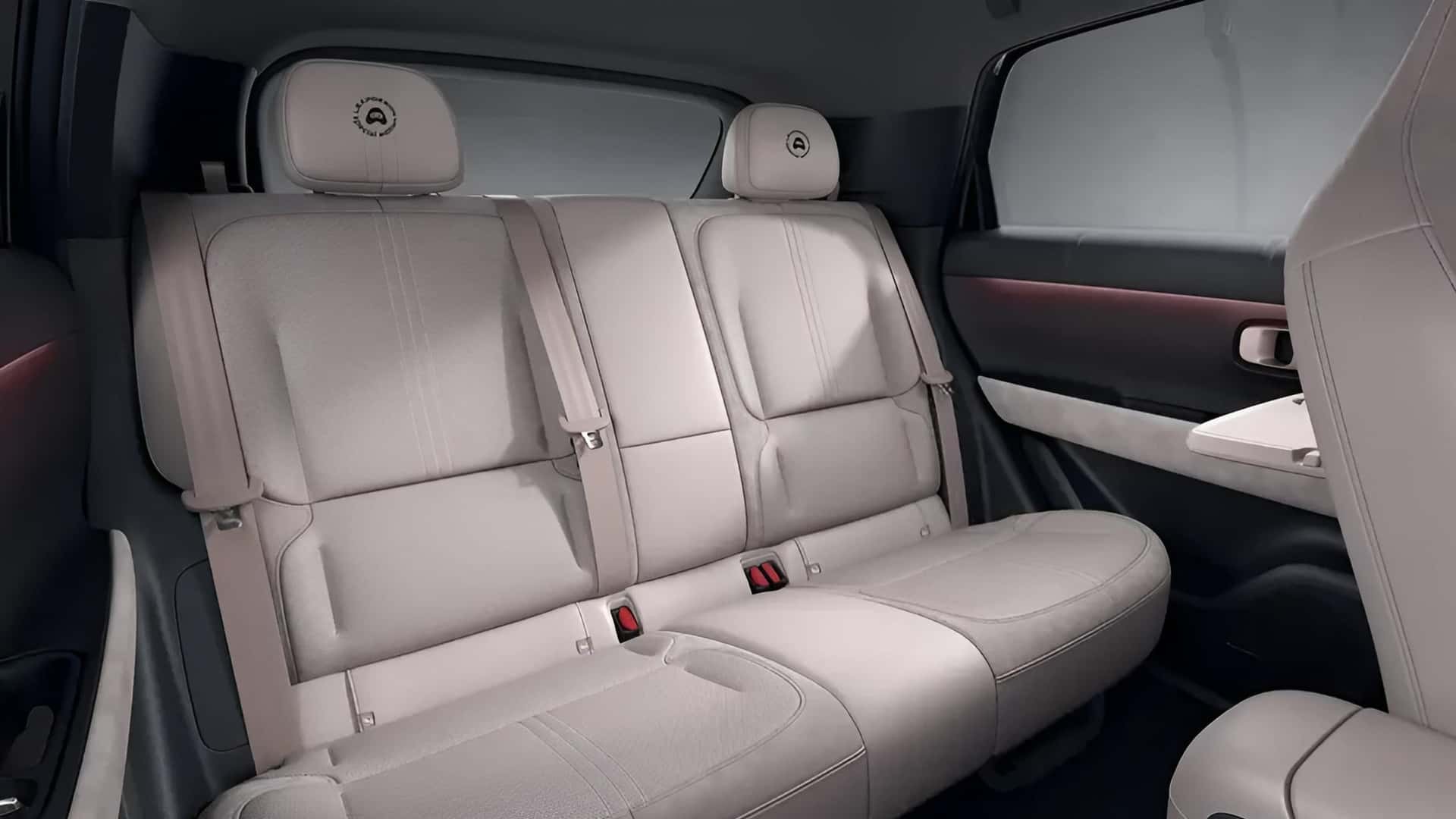 Interior de carro com bancos traseiros em couro bege claro, cintos de segurança visíveis e encostos de cabeça com um logotipo circular. A porta lateral tem detalhes em preto e uma faixa iluminada em vermelho, com uma pequena mesa dobrável no encosto do banco dianteiro
