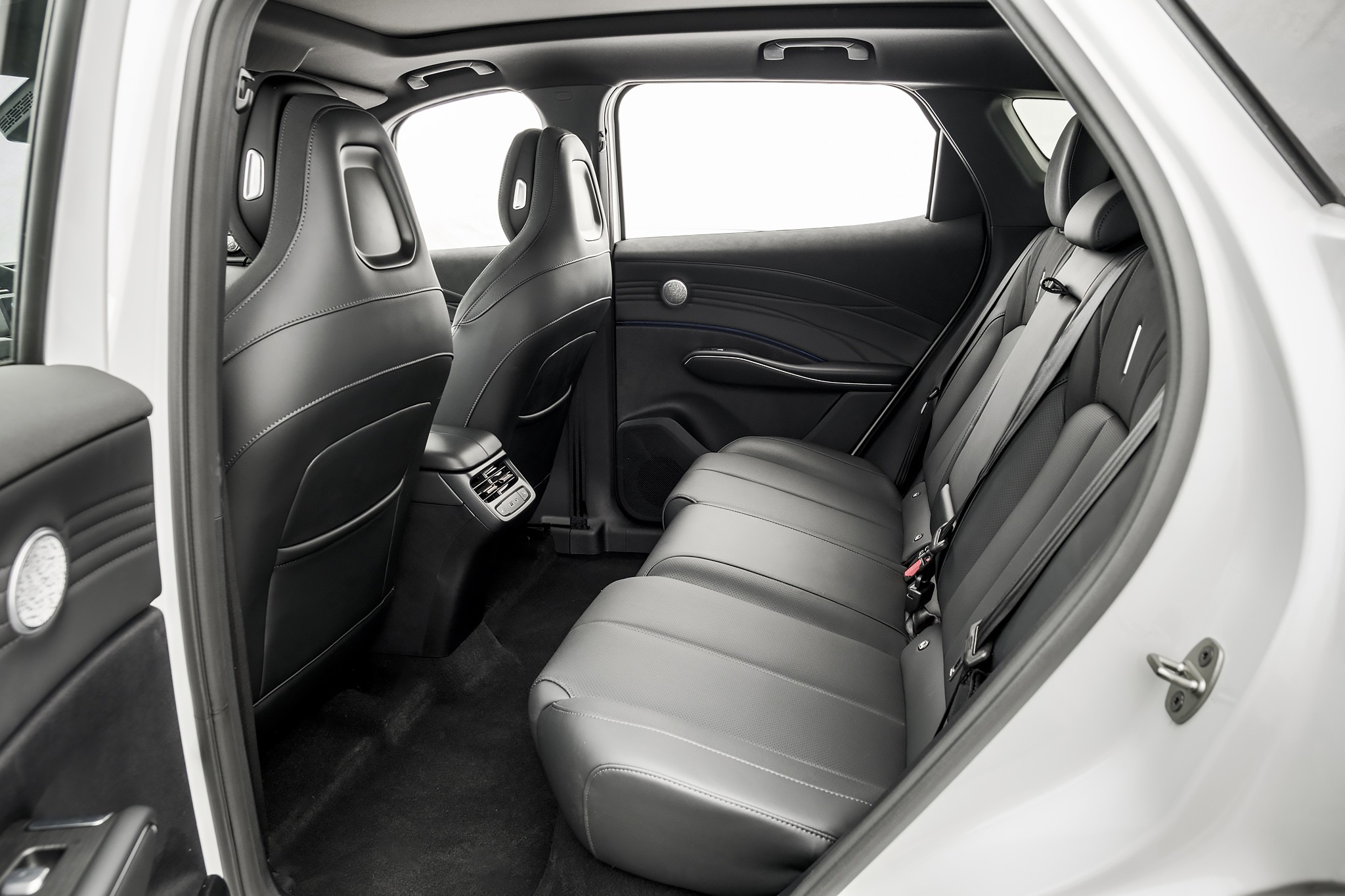 Interior de um carro, mostrando os bancos traseiros e a parte de trás dos bancos dianteiros, todos em couro preto com costuras brancas. O banco traseiro é inteiriço, com cintos de segurança visíveis. As portas têm acabamento preto e detalhes prateados. O teto solar e as janelas estão visíveis, com luz branca entrando. O piso é forrado com carpete escuro