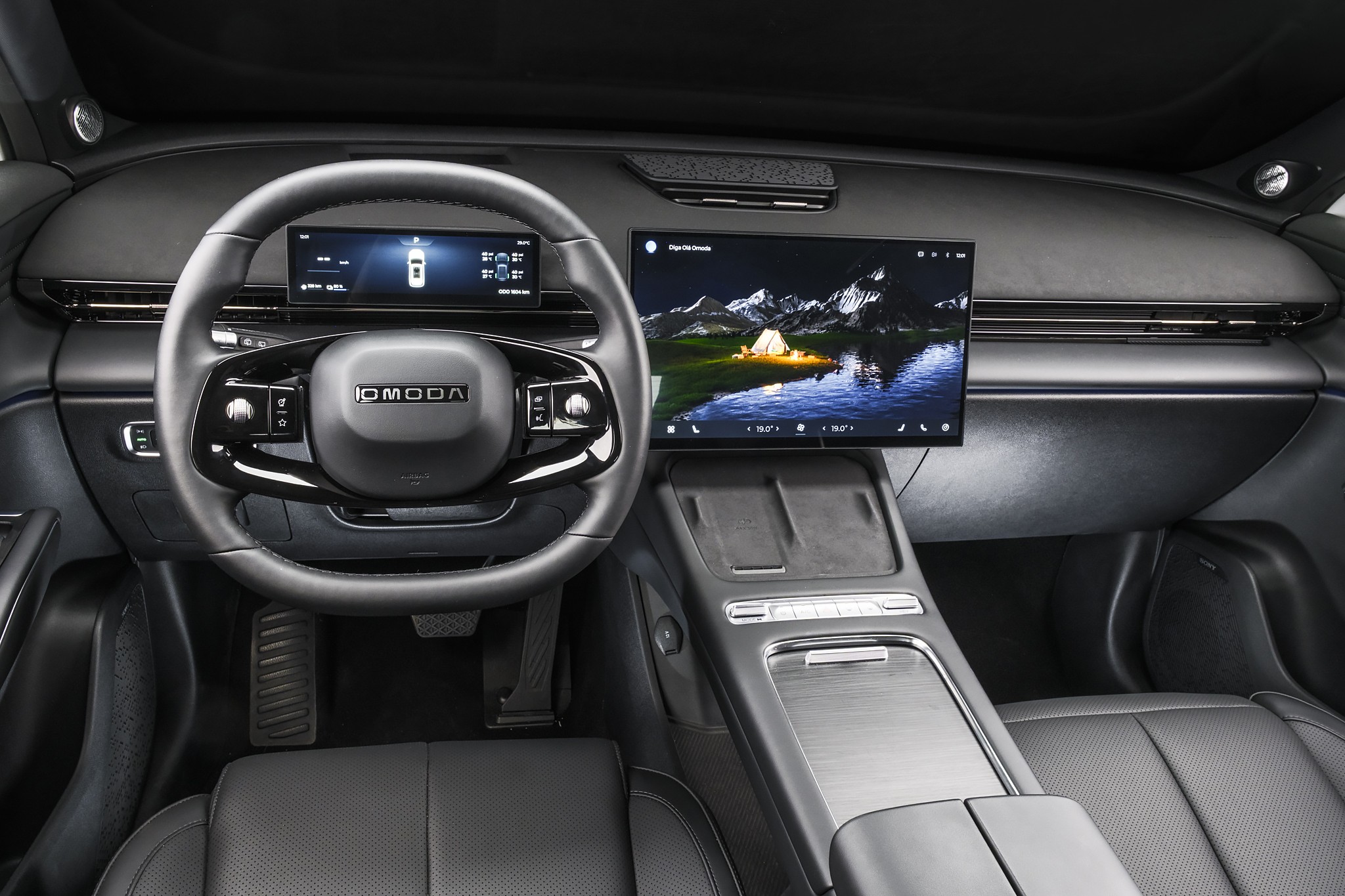 Interior de um carro moderno com volante preto multifuncional e logo OMODA. Dois displays digitais: um menor atrás do volante mostrando informações do veículo, e outro maior à direita exibindo uma paisagem noturna com montanhas, lago e uma barraca iluminada. O console central possui acabamento escuro e detalhes prateados, com bancos de couro pretos visíveis