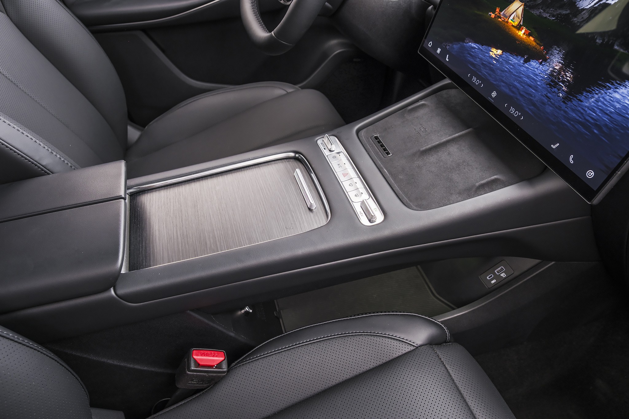 Interior de carro moderno com console central em preto e cinza escovado, bancos de couro perfurado e tela multimídia exibindo uma paisagem noturna com barco iluminado