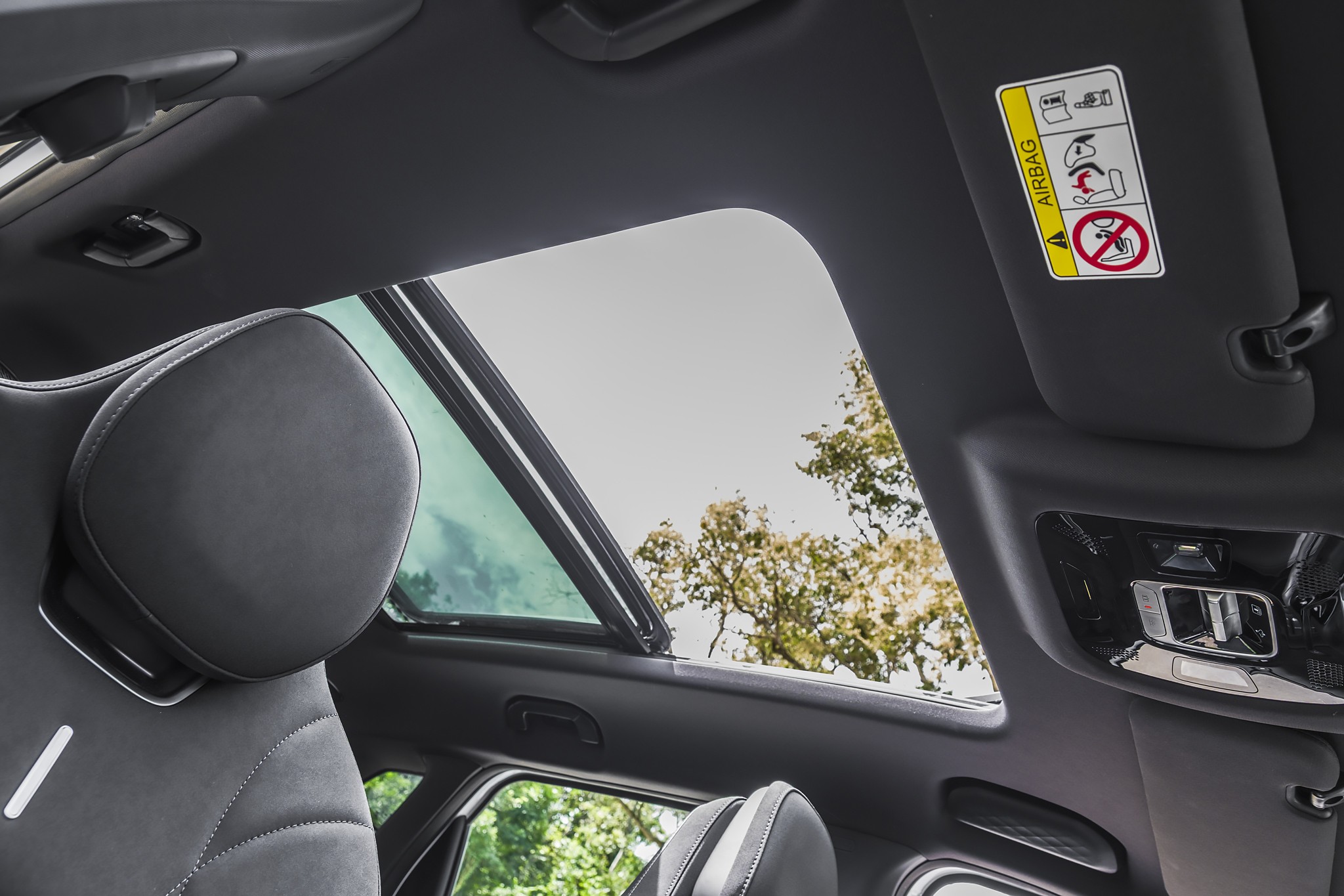 Interior de um carro com teto solar panorâmico aberto, mostrando o céu claro e árvores verdes. O encosto de cabeça do banco dianteiro esquerdo, estofado em couro preto com costuras brancas, está em primeiro plano. No para-sol direito, um adesivo de aviso de airbag é visível
