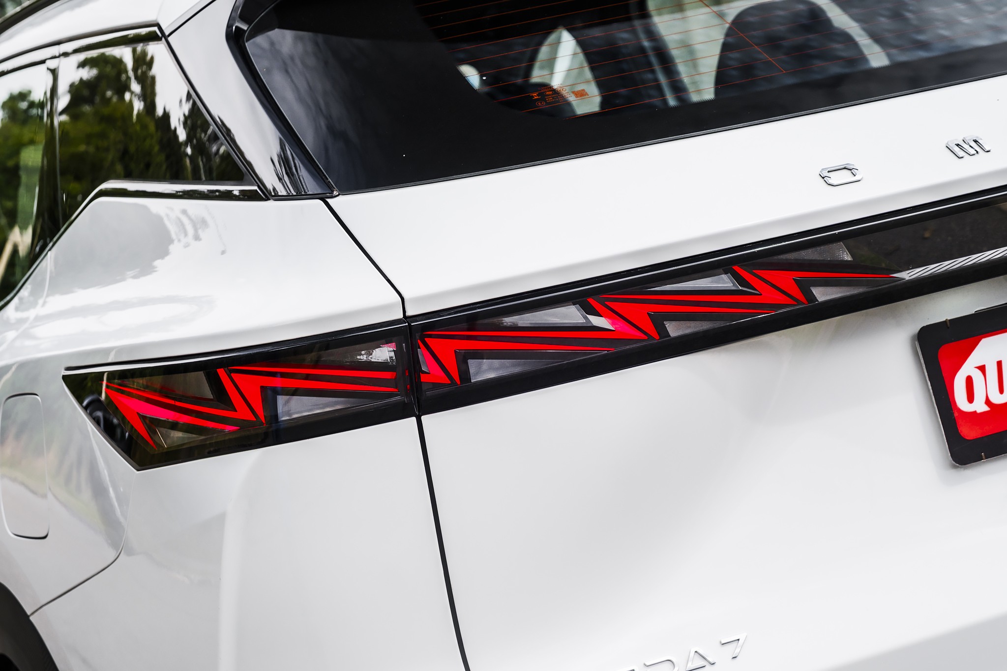 Lanterna traseira de LED vermelha e preta com design em zigue-zague, integrada à carroceria branca de um carro, com reflexos de árvores no vidro traseiro