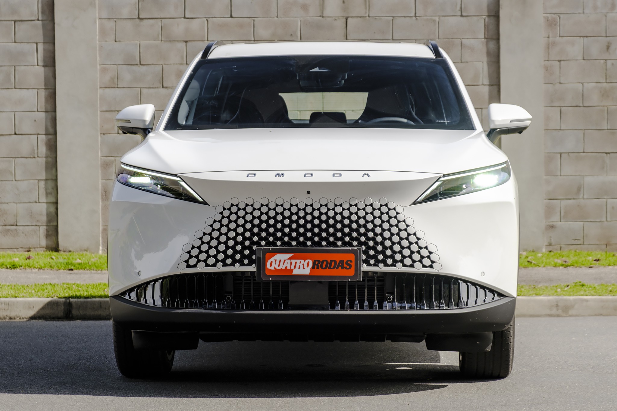 Carro branco Omoda de frente, com grade frontal em padrão de colmeia e faróis finos acesos, estacionado em rua asfaltada com parede de tijolos ao fundo. Placa QUATRO RODAS no centro da grade.