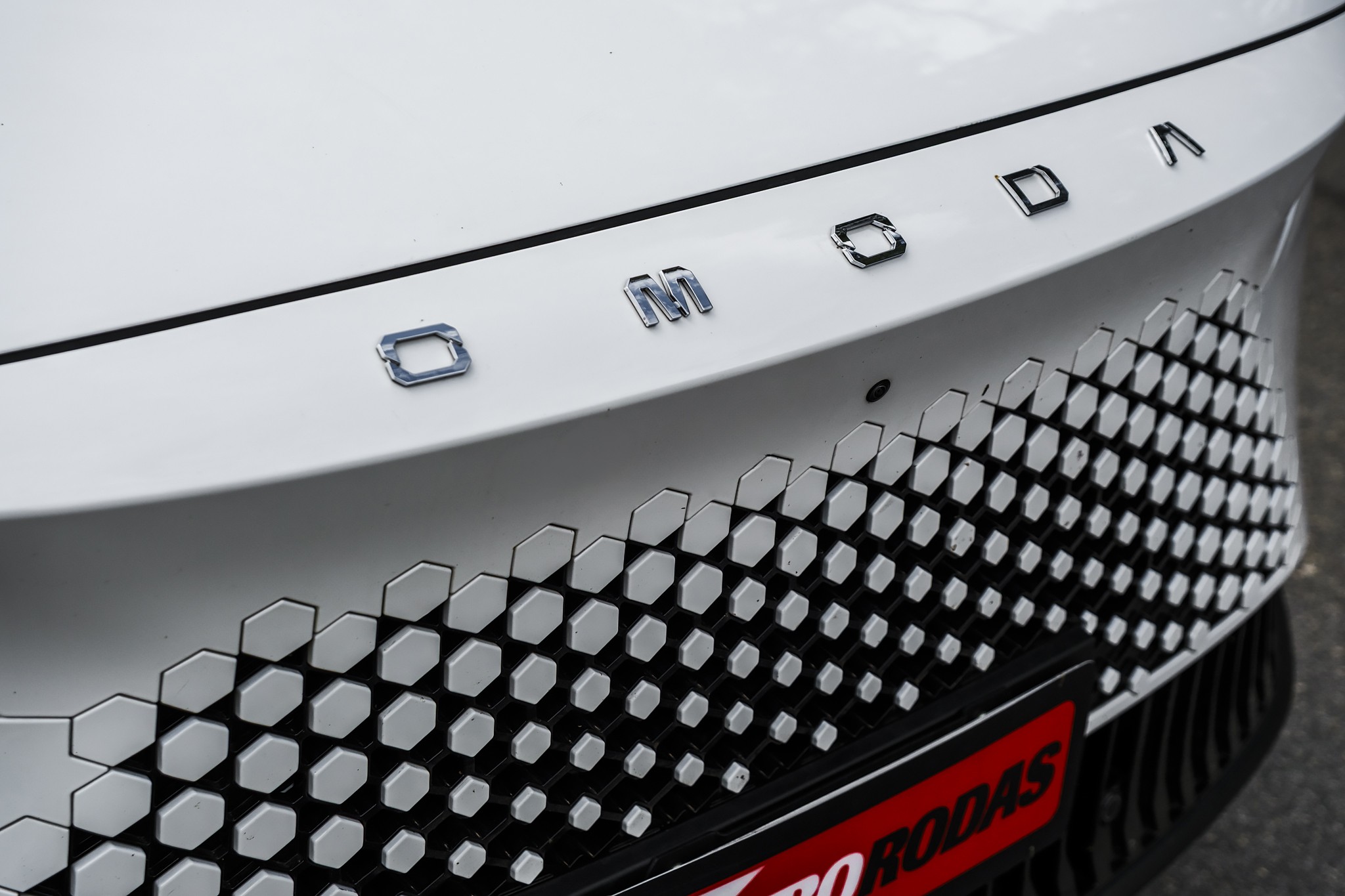 Traseira de carro branco com o nome OMODA em letras cromadas e uma grade inferior preta com padrão de hexágonos brancos e cinzas. Uma faixa vermelha com RODAS em branco aparece na parte inferior