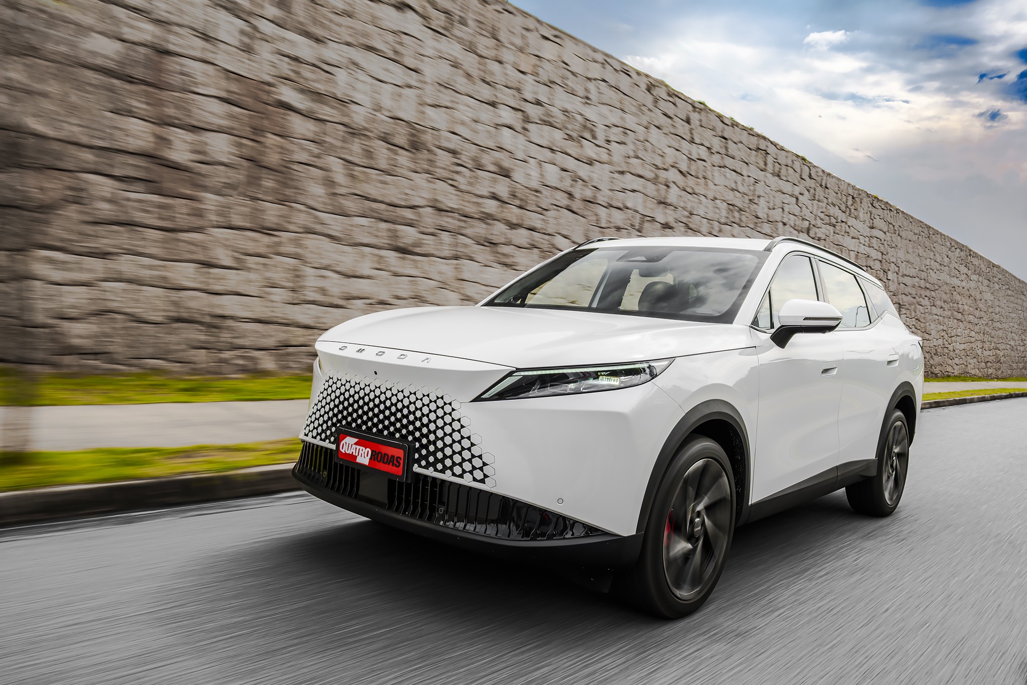 SUV branco moderno em movimento, visto de frente e lateral, com grade frontal em padrão hexagonal e placa vermelha Auto Esporte. Ao fundo, um muro de pedras e céu nublado, com asfalto borrado indicando velocidade