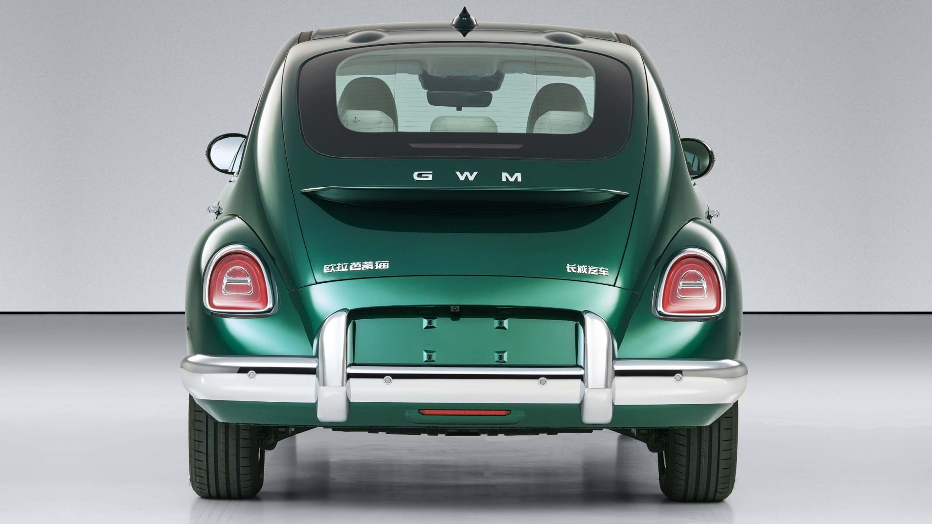Traseira de um carro verde-escuro com design retrô, similar a um Fusca, em um estúdio cinza. O carro tem para-choques cromados, lanternas traseiras ovais vermelhas e o logo GWM no porta-malas. Pneus pretos com aros prateados completam o visual.