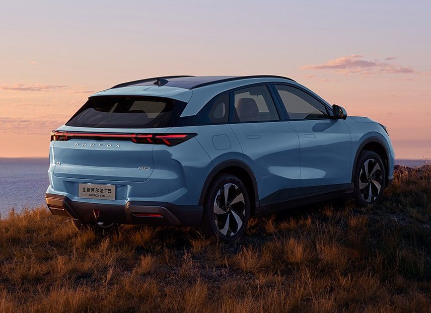 SUV azul claro estacionado em uma colina gramada com vista para o oceano ao pôr do sol, céu em tons de laranja e rosa