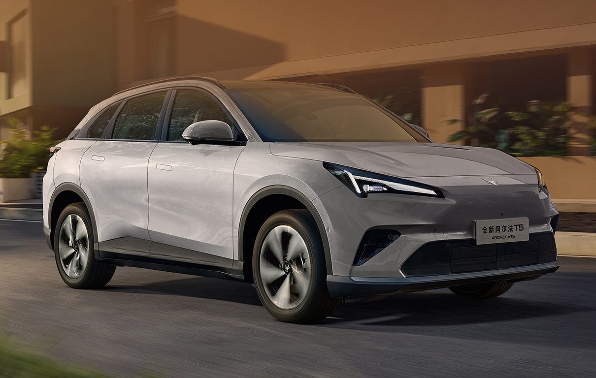 SUV elétrico cinza claro com detalhes escuros na frente, rodando em uma rua asfaltada com prédios modernos e árvores ao fundo, sob luz solar