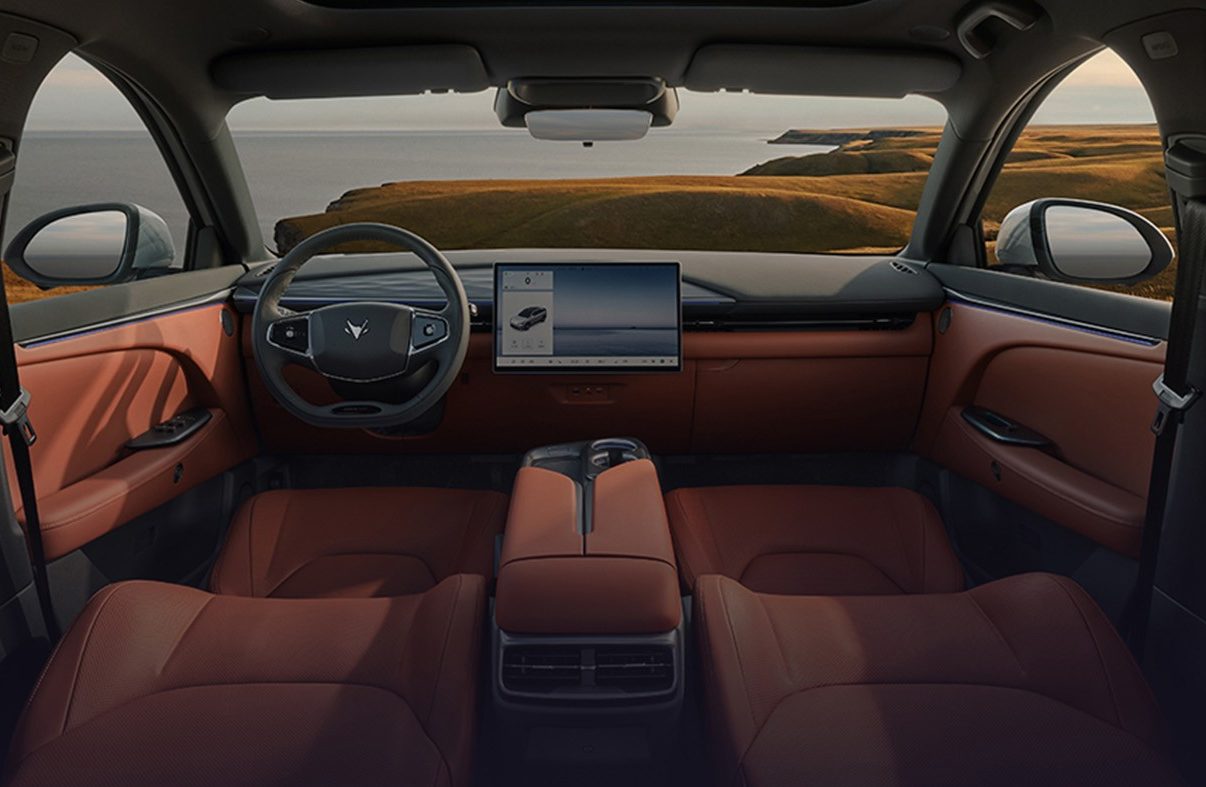 Interior de carro moderno com bancos e painéis de porta em couro marrom, teto solar panorâmico e tela multimídia. Ao fundo, paisagem costeira com mar e colinas gramadas sob céu azul com nuvens esparsas