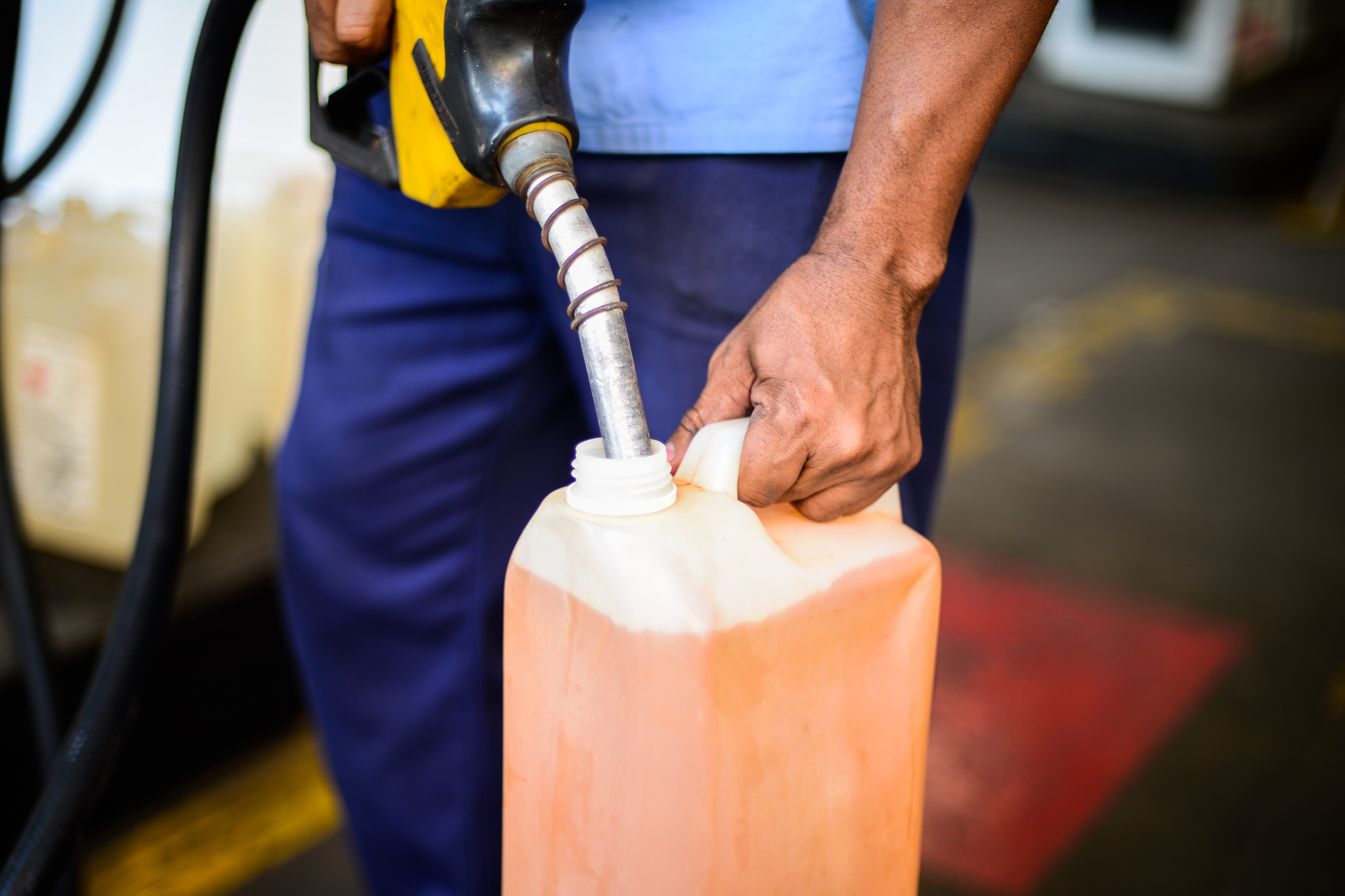 Frentista com uniforme azul, segurando um bico de bomba de combustível amarelo e preto, abastecendo um galão plástico laranja com líquido da mesma cor