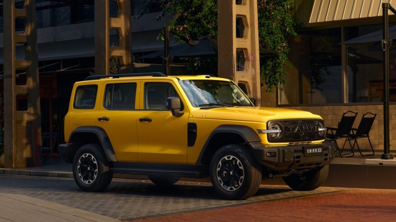 SUV amarelo vibrante, estilo off-road, estacionado em uma rua urbana com prédios e folhagem ao fundo. O veículo tem teto preto, para-lamas robustos e rodas escuras com detalhes prateados. Duas cadeiras pretas dobráveis são visíveis à direita.