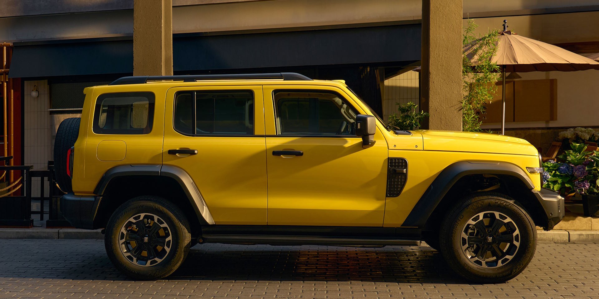 SUV amarelo vibrante estacionado na rua, com pneu sobressalente na traseira e detalhes em preto fosco. Ao fundo, um prédio e um guarda-sol bege.