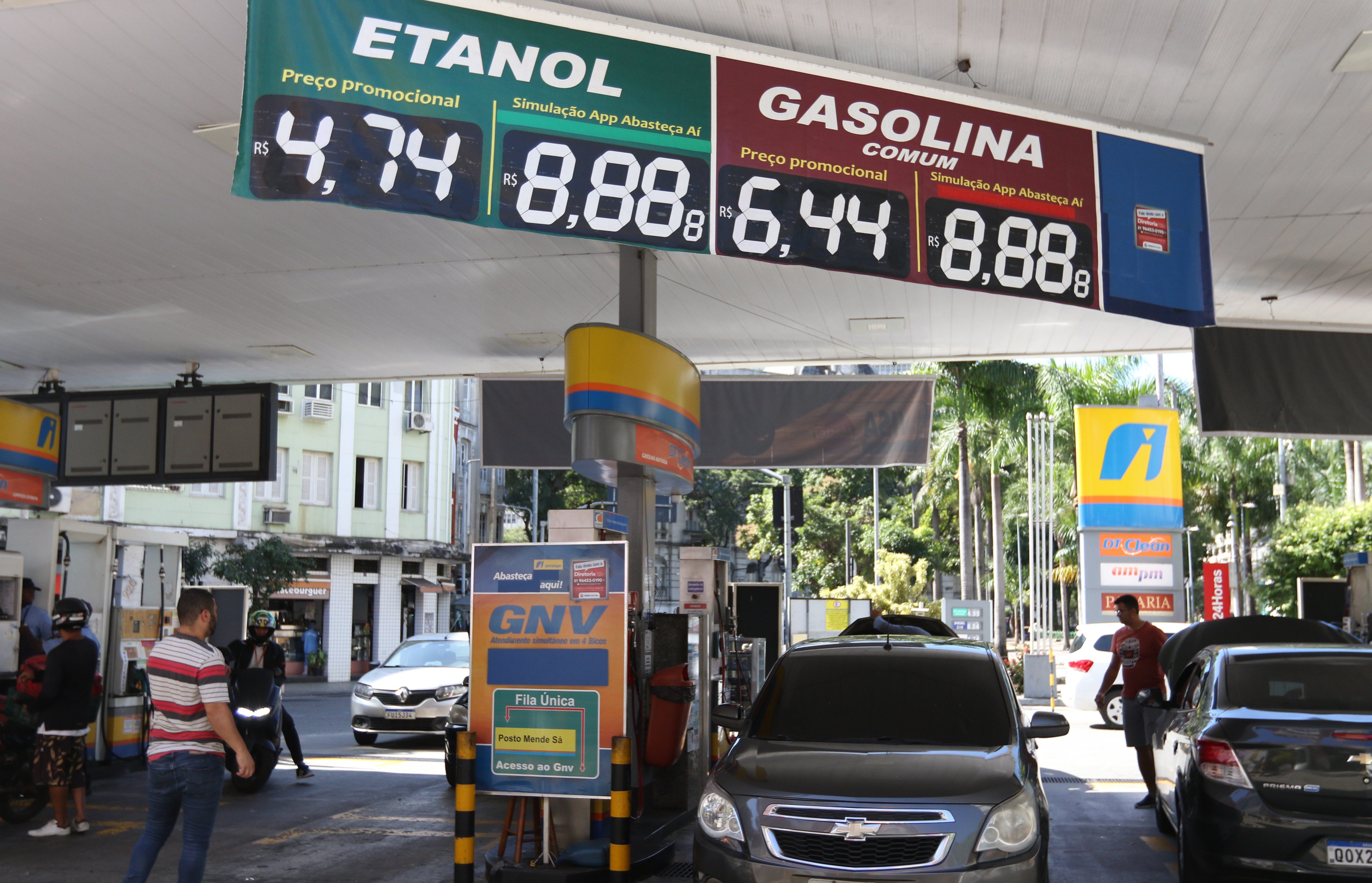 Posto de gasolina com letreiros digitais exibindo preços: Etanol a R$ 4,74 e Gasolina Comum a R$ 6,44. Carros e pessoas circulam no local, com um painel de GNV à direita e prédios ao fundo