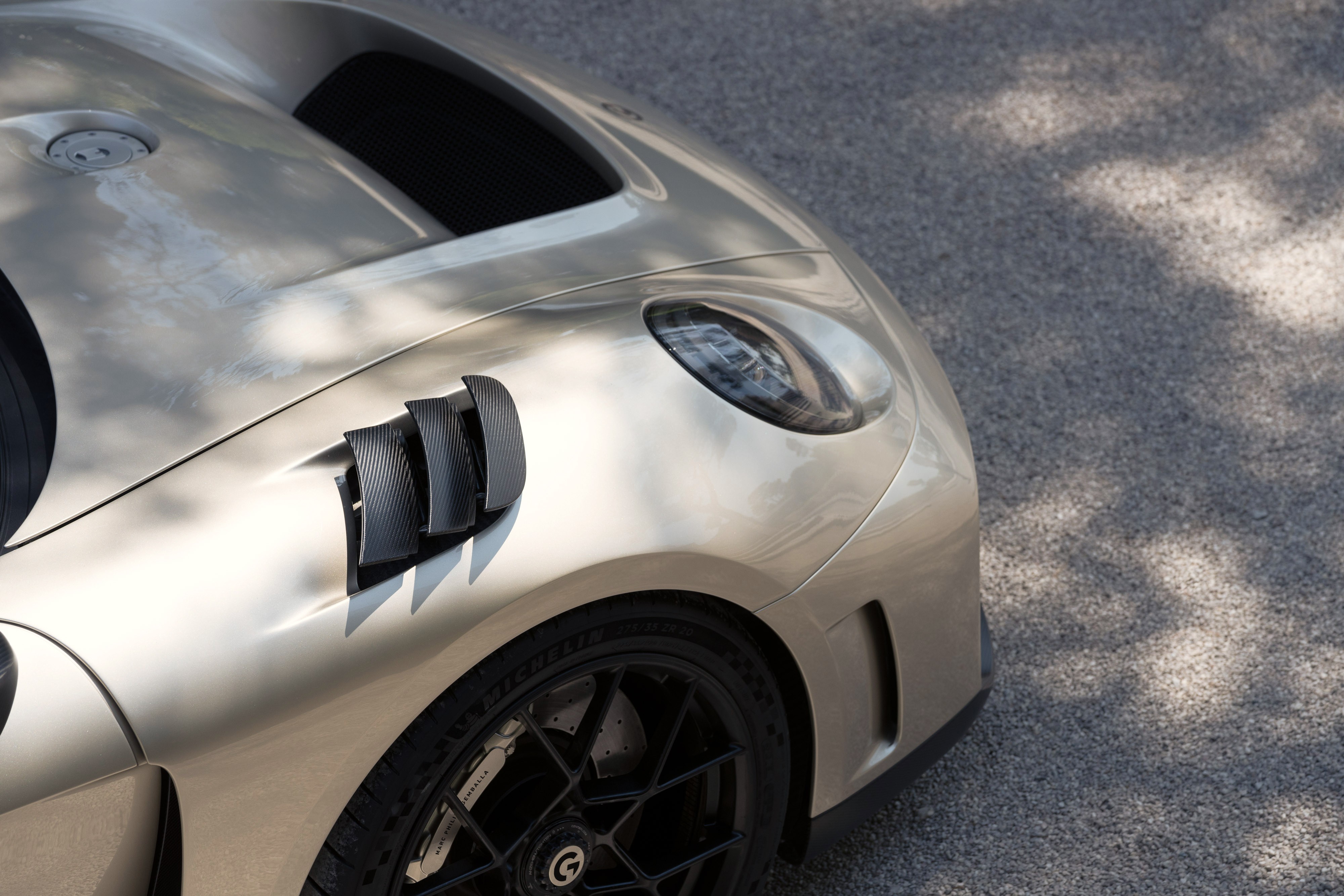 Lateral de um carro esportivo dourado ou champanhe, com três aberturas de ventilação pretas e farol oval. A roda preta com design geométrico é visível na parte inferior, sobre asfalto cinza com sombras de árvores